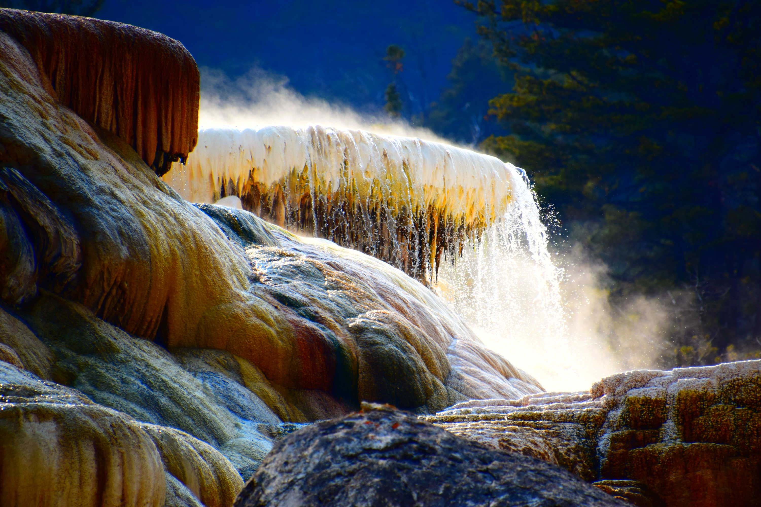 Evaporation catches sunlight behind a backlit Mammoth Hot Springs Terraces, leading to a stark and saturated bright outline. Steamy water cascades down the terrace.