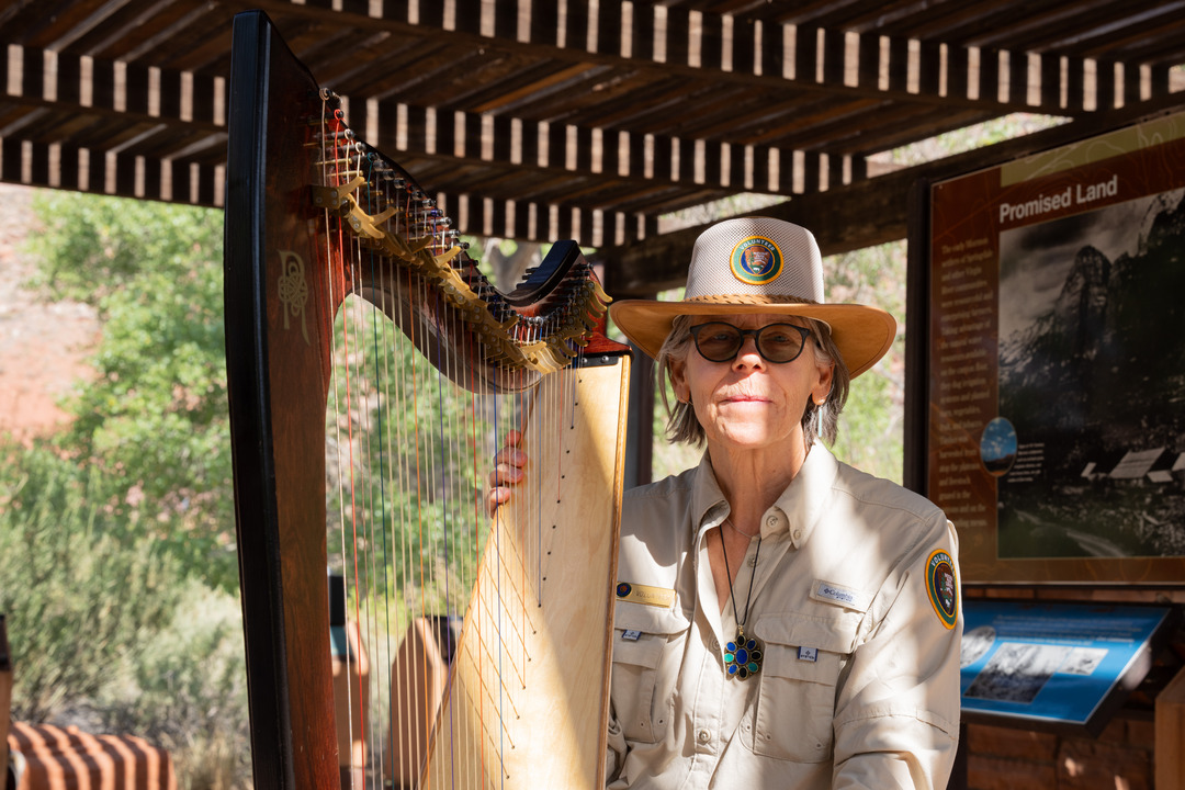A woman in a National Park Service volunteer uniform sits next to a large, wooden harp. Overhead, a wooden pergola casts shade across the two.