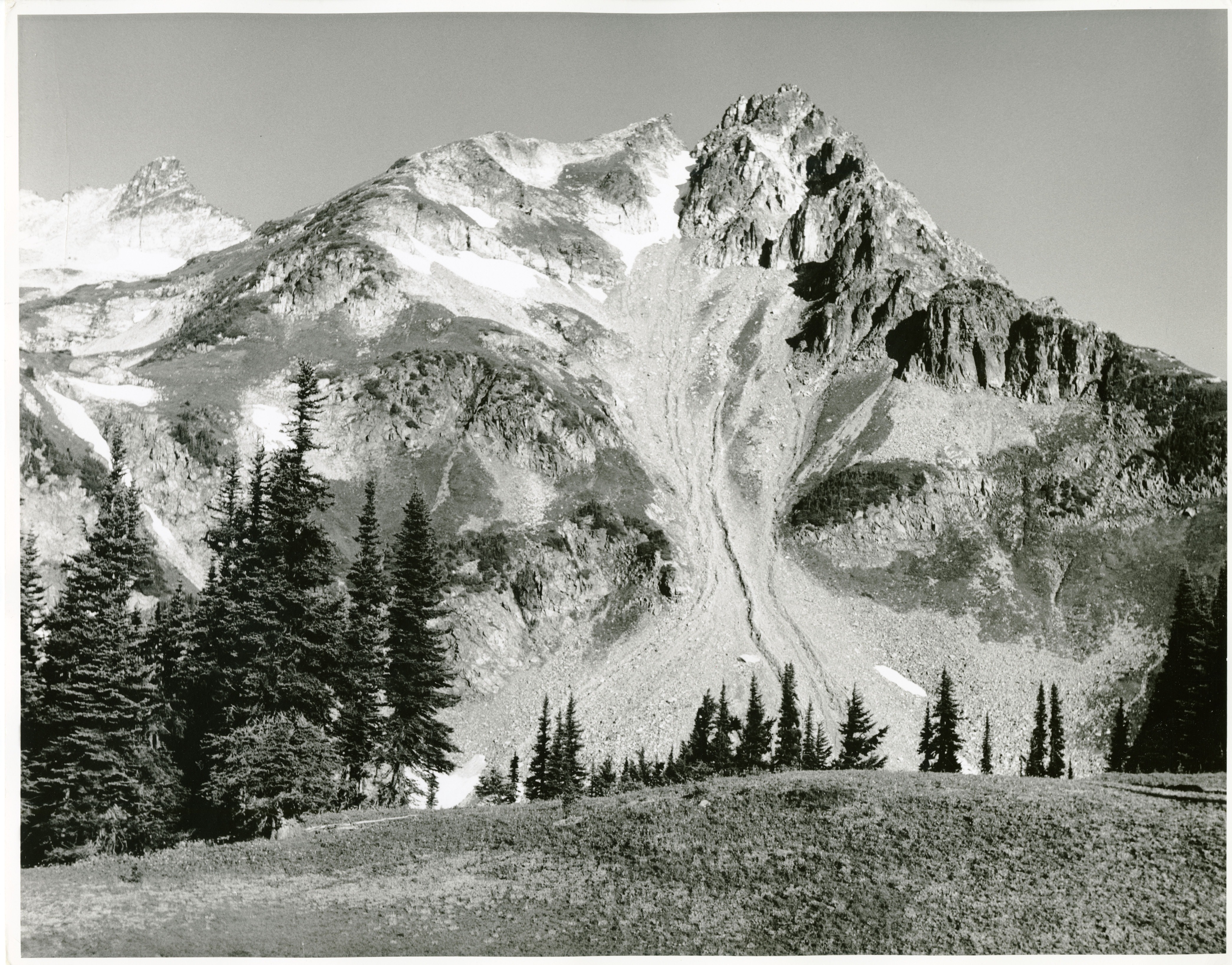 A rocky mountain in front of a grassy slope. Evidence of heavy erosion on the mountain. Coniferous trees between the slope and the mountain. (North Cascades NPS Complex Museum Collection, Assembled Collections, Series V: NPS North Cascades Survey Photographs)