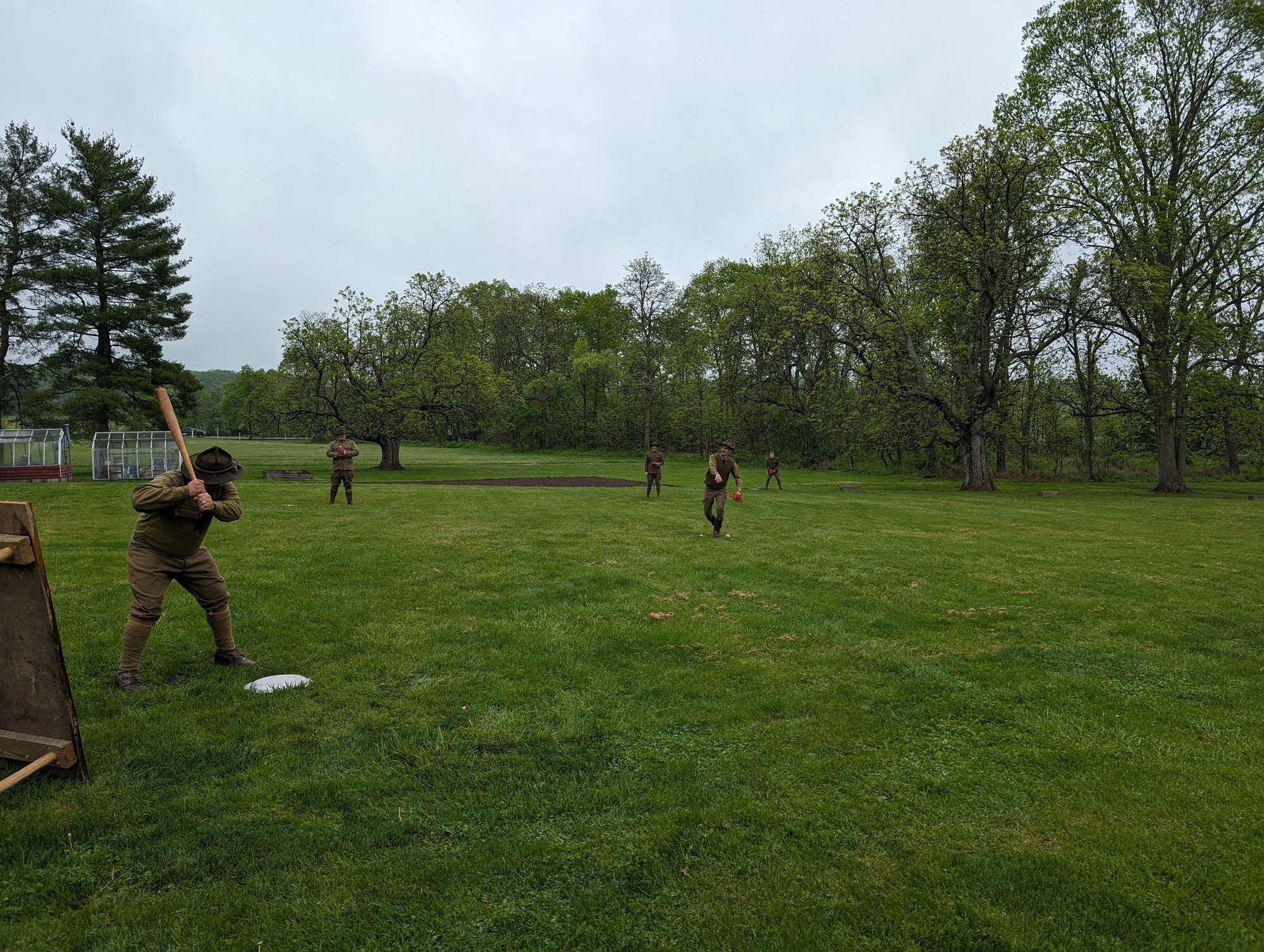 A group of living historians in military uniforms play baseball on a grassy field near trees. 