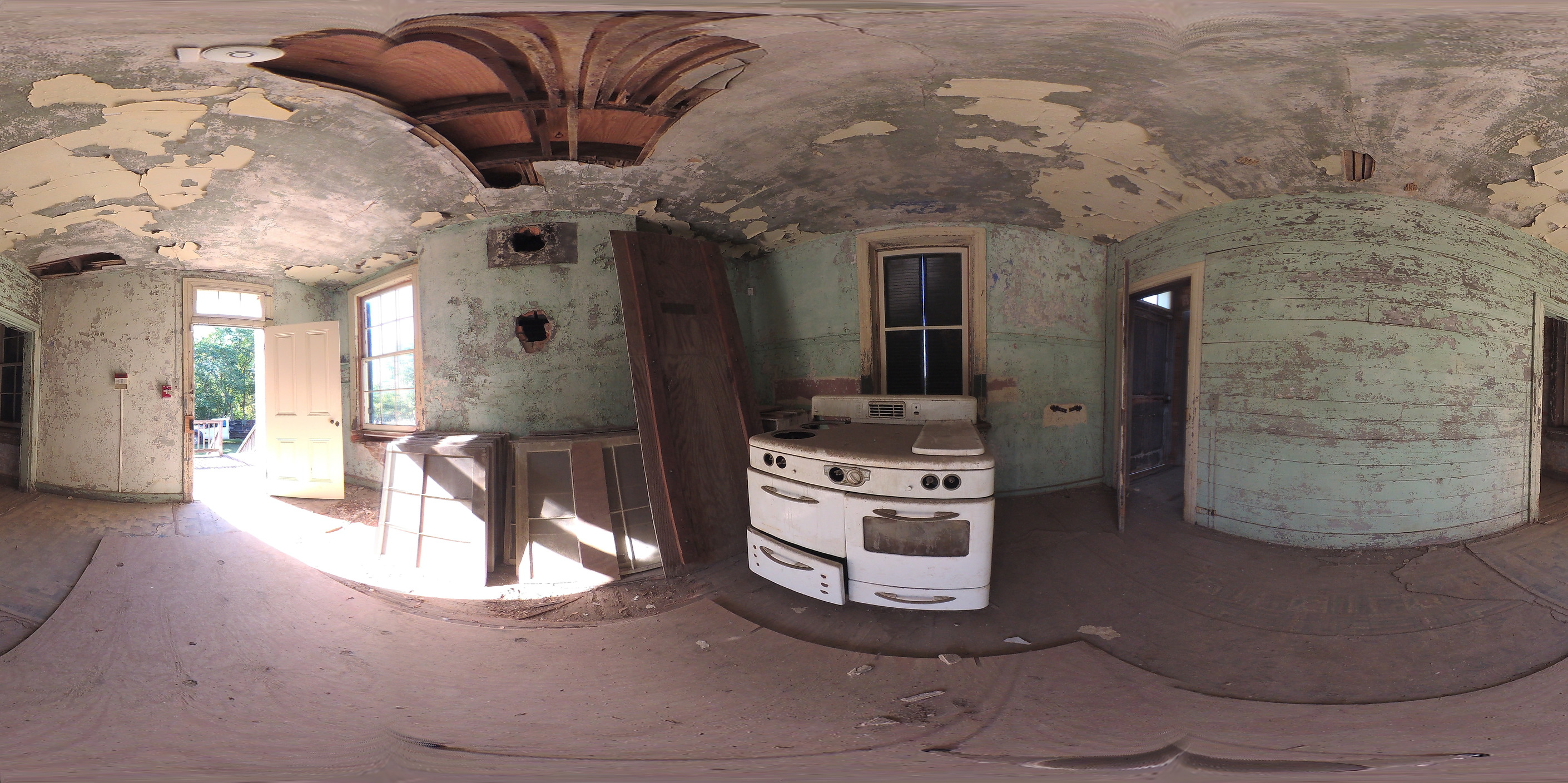 An open door allows sunlight to fill the medium-sized room. On the wall adjacent to the door, a doorway to the left leads to the servants’ stairs, while a doorway to the right leads to the dining room.  About a dozen glass window panes line the right side of the kitchen floor. A stove sits opposite the open door. 