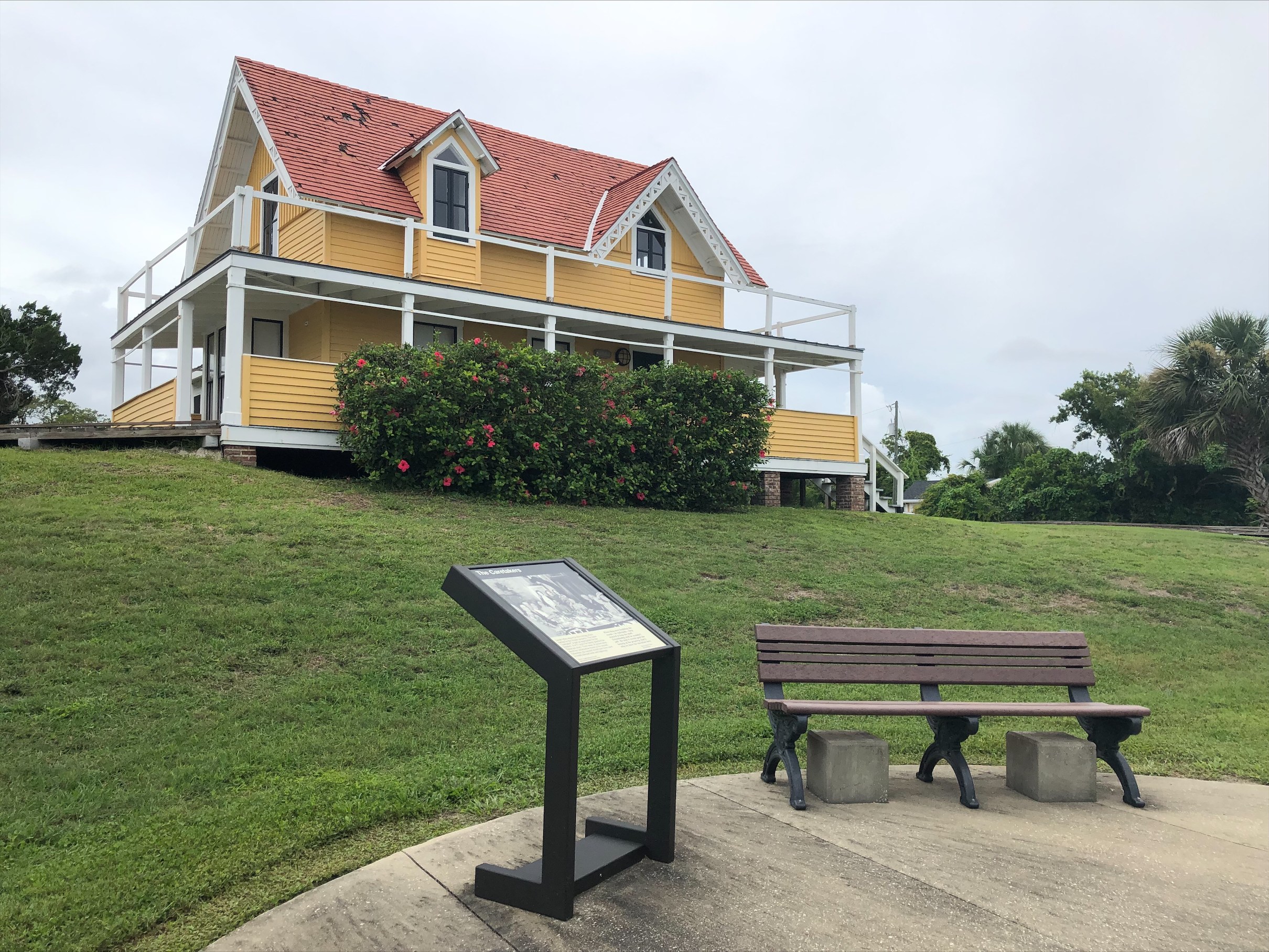 The cement sidewalk has an informational sign and a bench. In the background is the caretaker's house which is a gothic revival style home. 