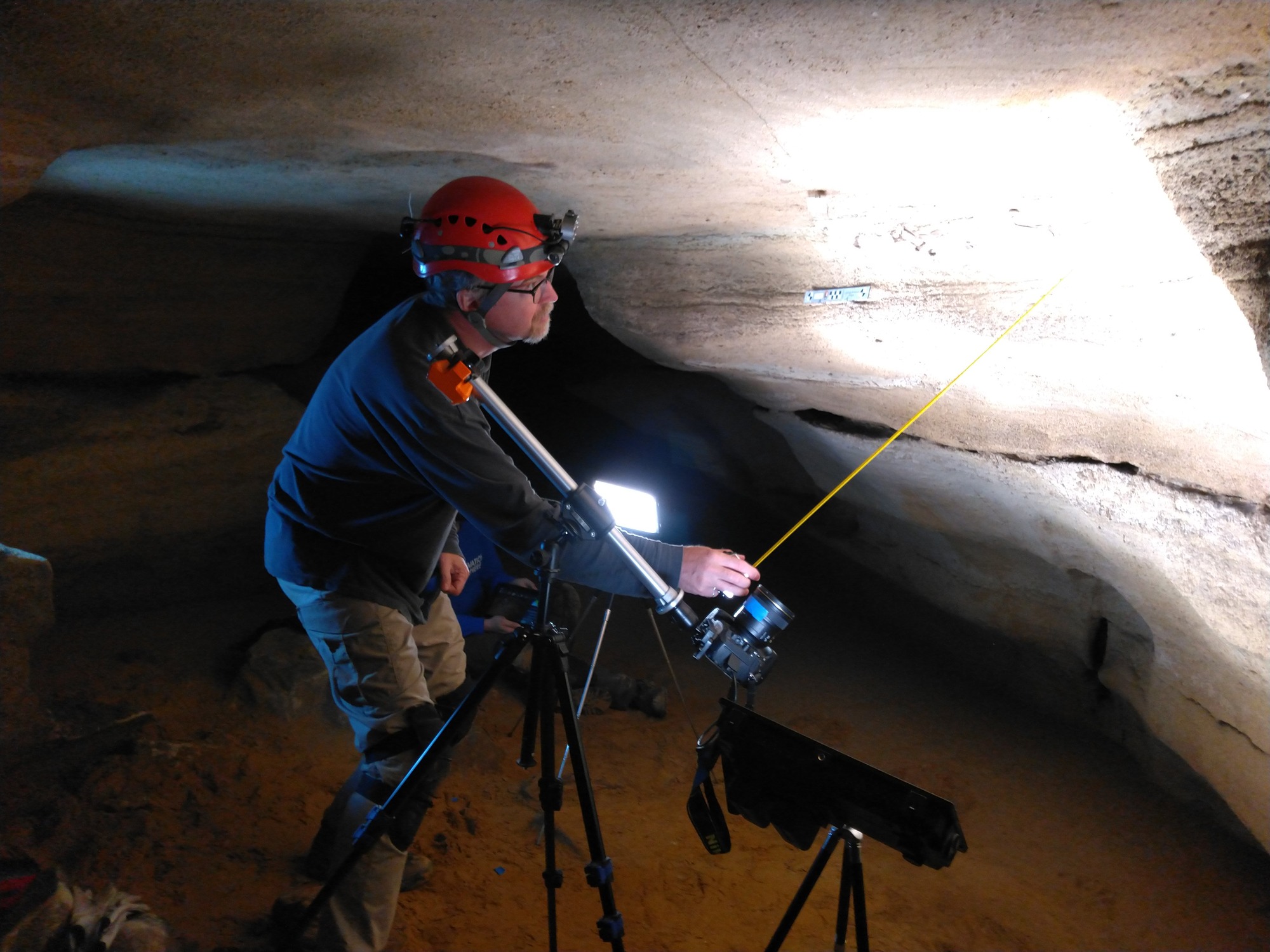 A man uses a yellow stick to determine the aim of a camera lens at a cave wall.