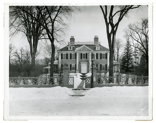 Facade of Georgian mansion in snow, surrounded by fence. Black and white photo.