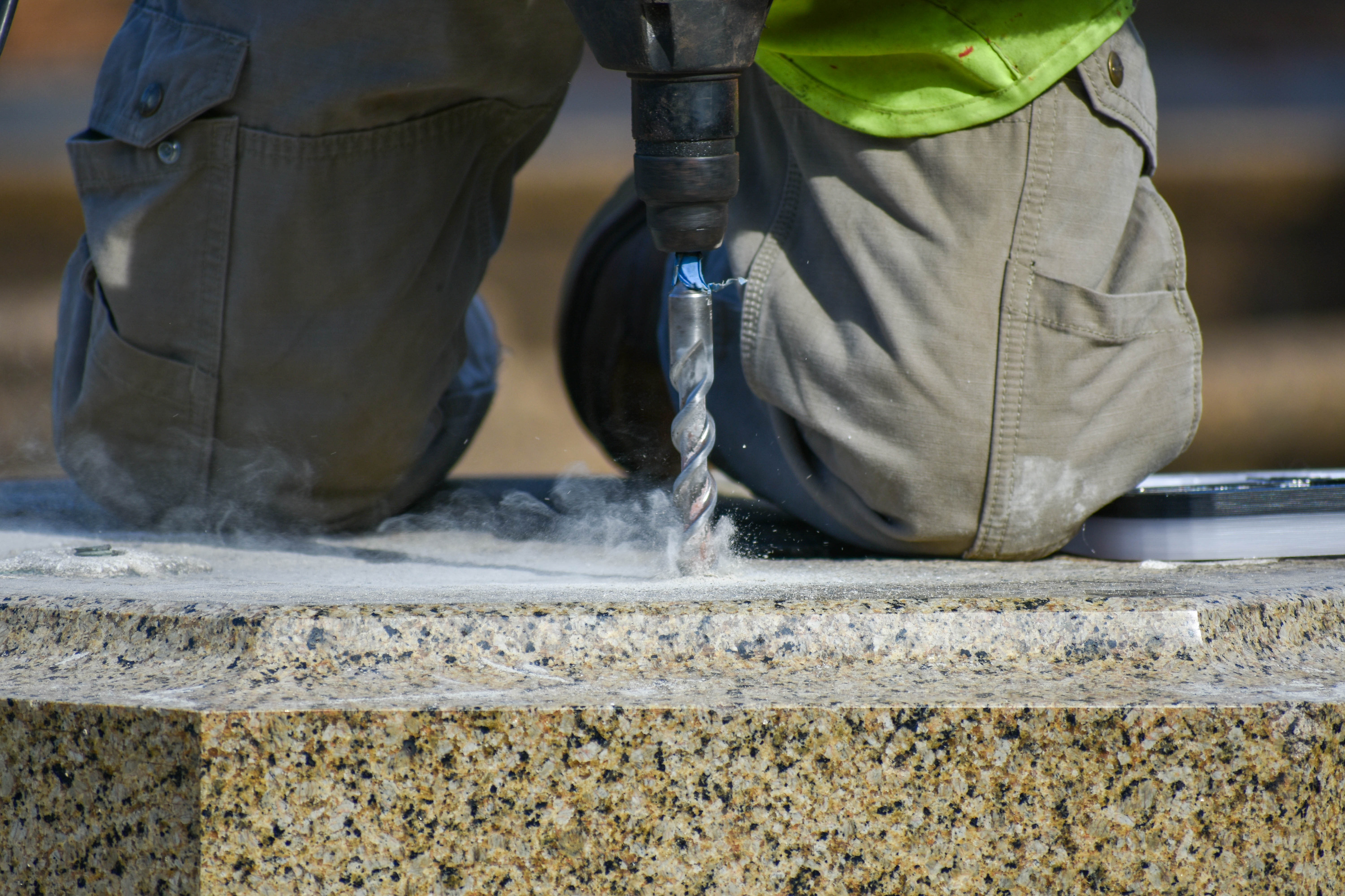 A close-up photo of a man on his knees drilling into a granite platform. Fine particles fly up from the drill and clouds of dust surround it.