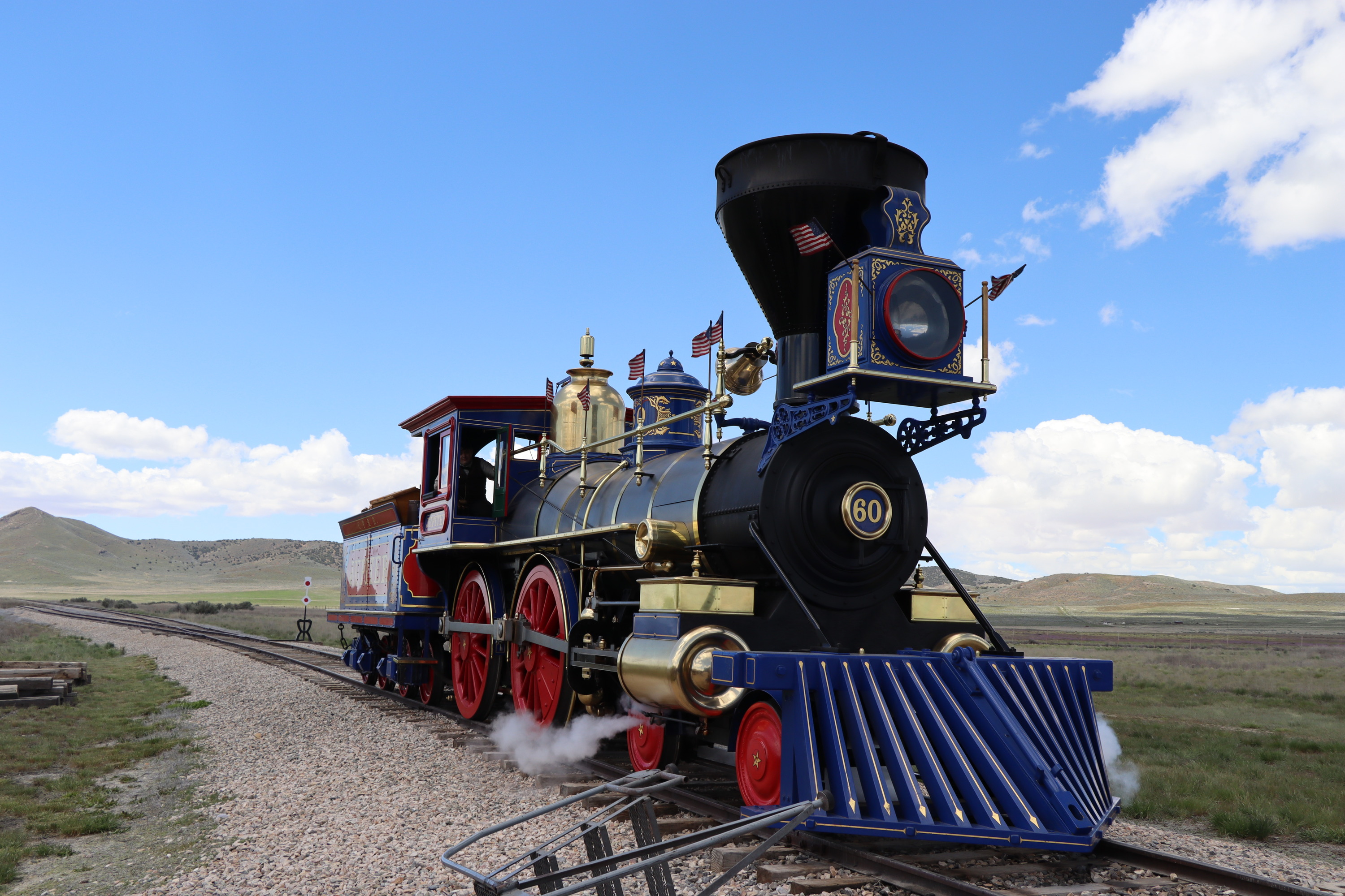 The blue, red, and gold steam engine Jupiter sits on railroad tracks. White steam billows from the wheels, matching the white clouds in the blue sky.

