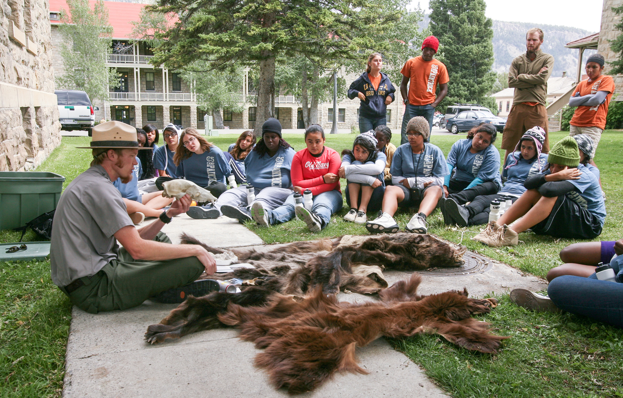 Male ranger sits on a sidewalk with animal pelts and talks with a large group of teenager, many of which are also seated on the ground.