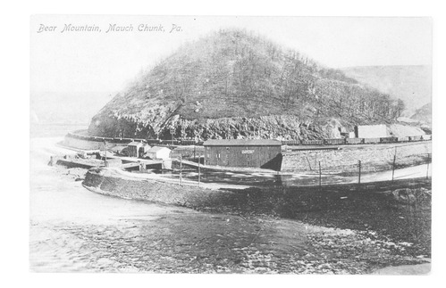 Postcard. "Bear Mountain, Mauch Chunk, PA." top left. Photograph of coal train encircling mountain. "For Comfort and/Economy Use a [Illegible] Stove" sign on shed, center. River in foreground. Reverse: "No. 1075D. Pub. By A.H. Luckenbach. Made in Germany" vertically at left.