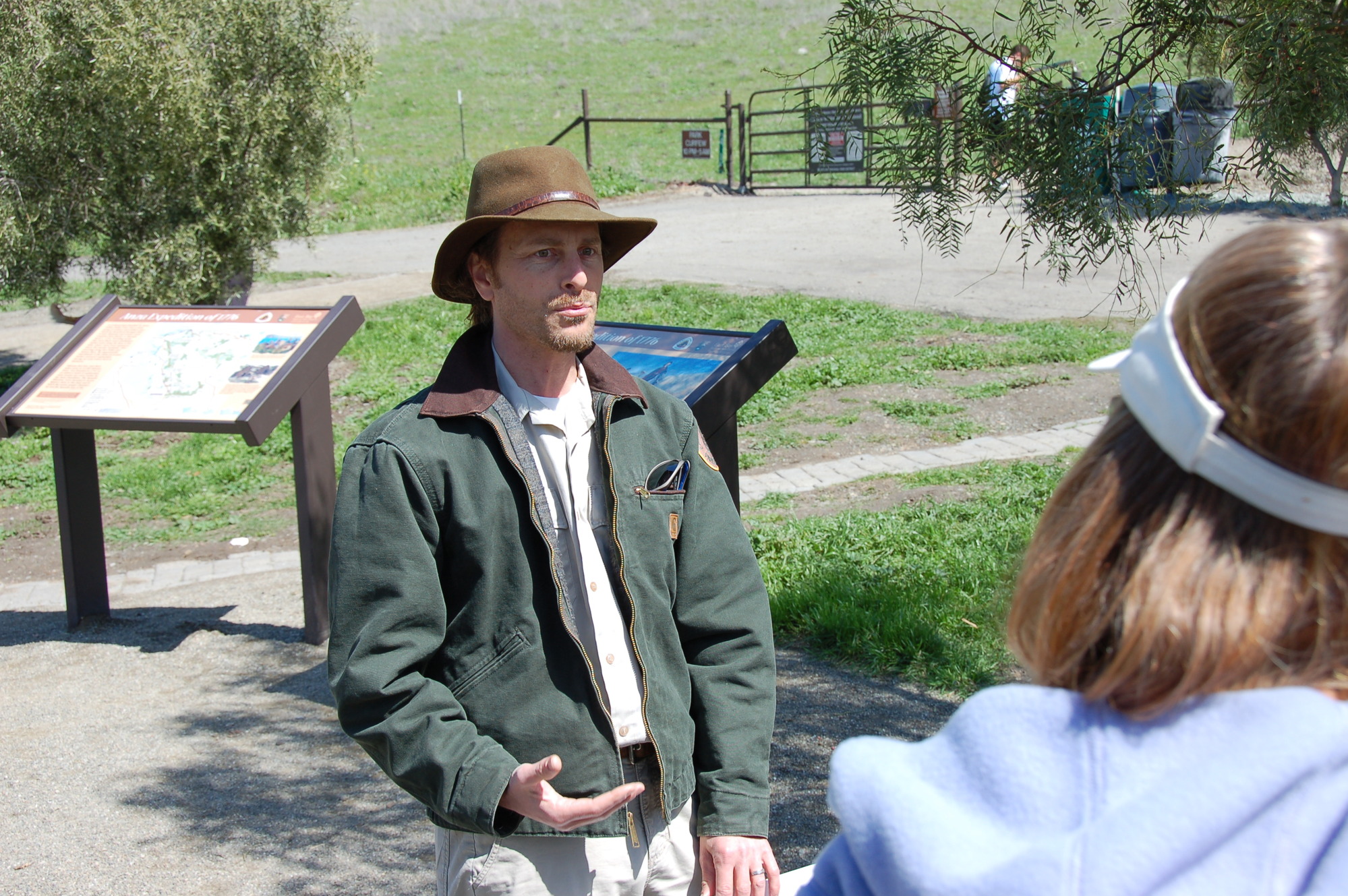A person wearing NPS uniform stands in front of waysides and speaks to a person with shoulder-length hair and a visor
