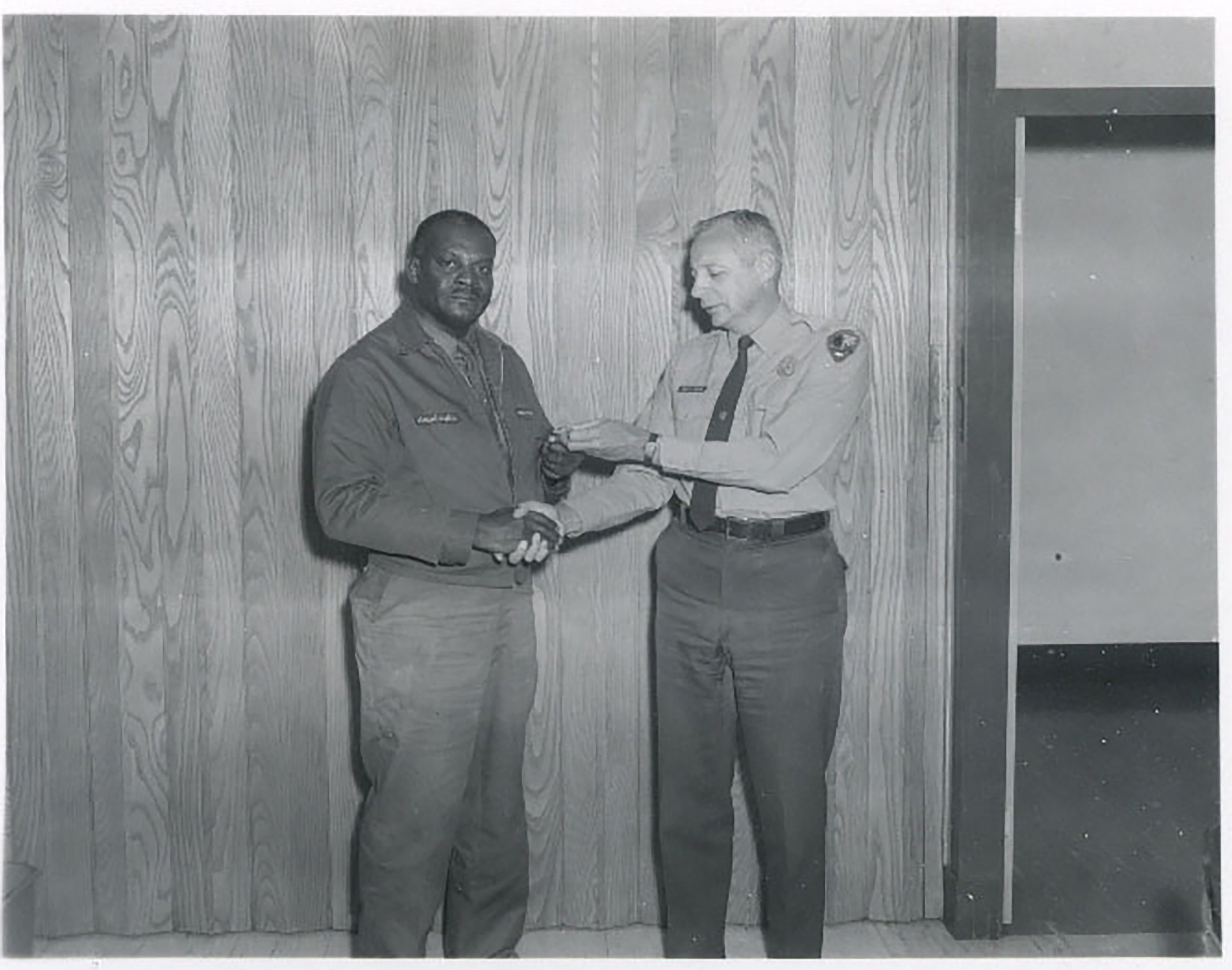An African American man in maintenance overalls, shakes hands with a Whtie man in a Ranger uniform. who is holding some sort of medal. 