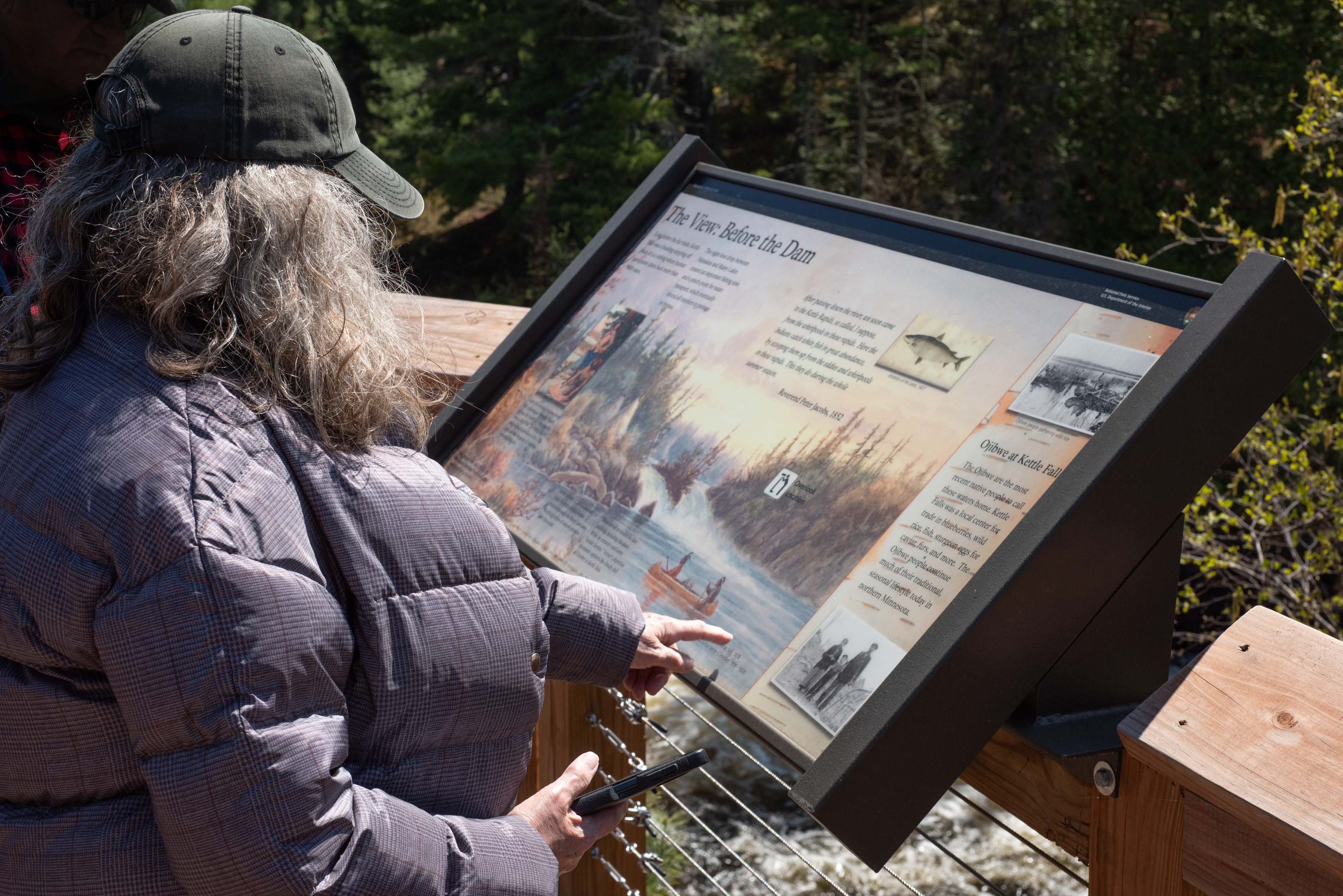 Photograph of person pointing at wayside sign with information relating to Kettle Falls. 