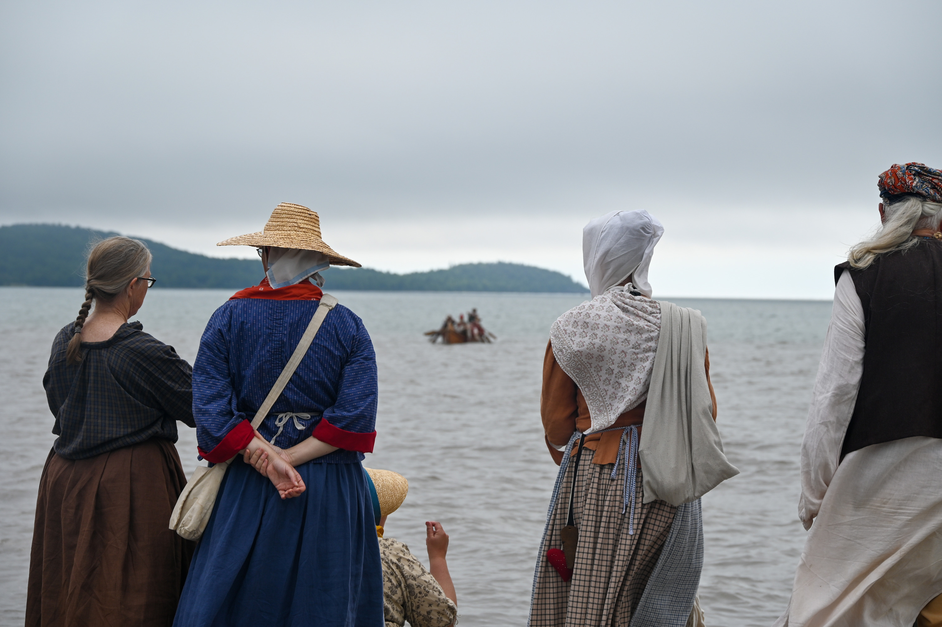 Five people in historic clothing face the water as a large canoe approaches.