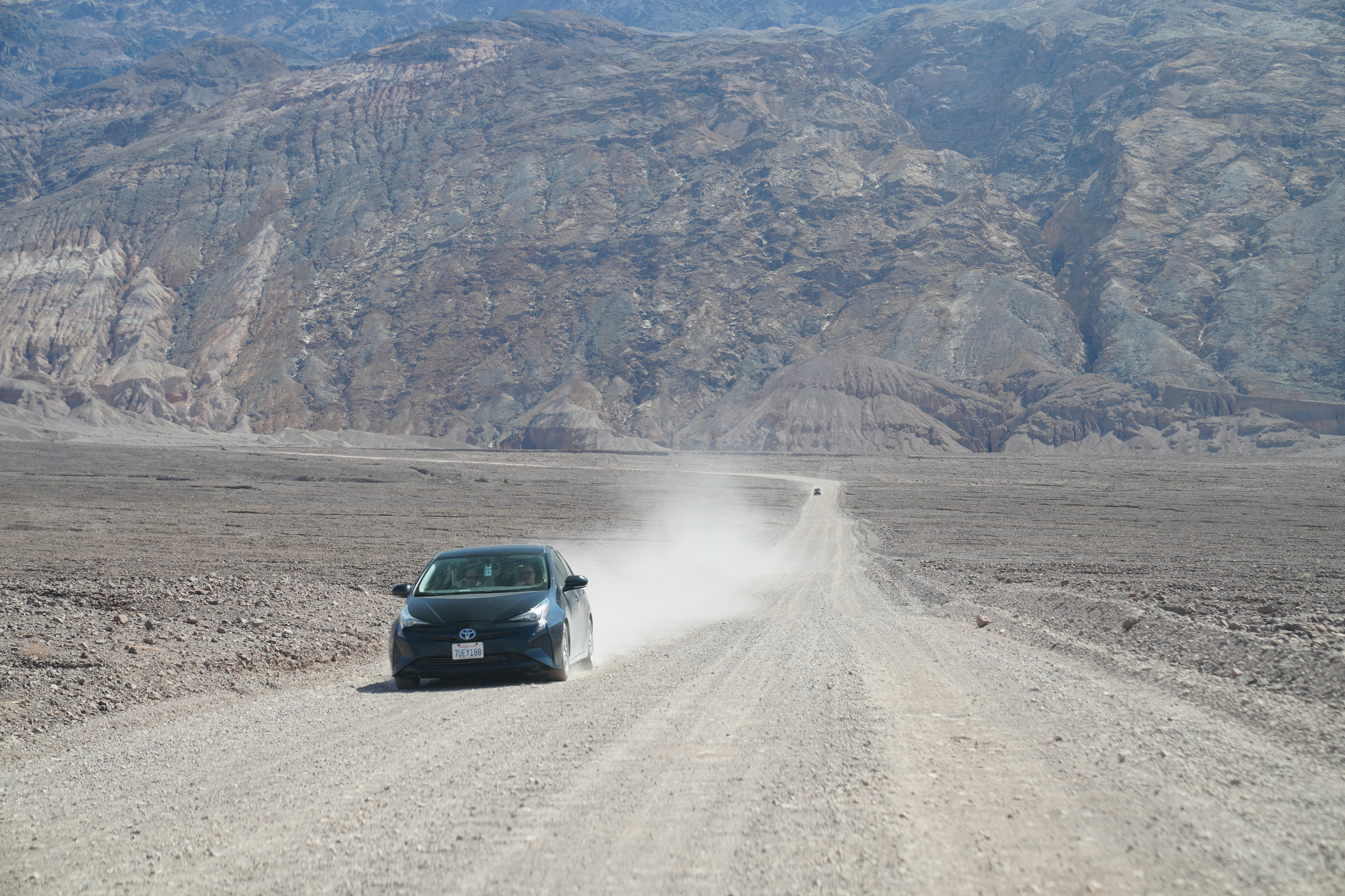 Natural Bridge road. A prius drives down a gravel road with a dust cloud trailing behind it. The road curves up and towards the base of the mountains in the background.