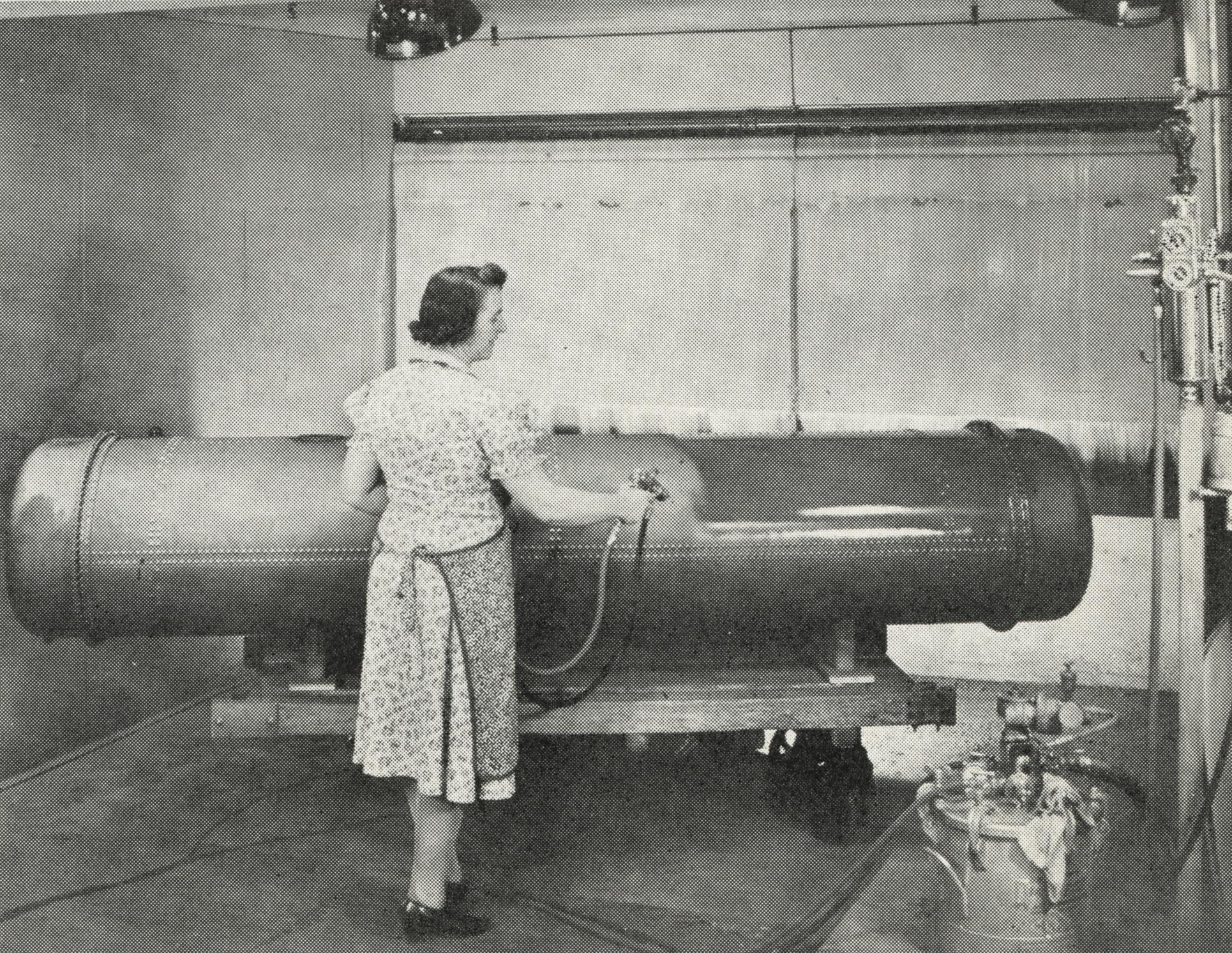 Black and white photo of the back of a woman standing wearing a dress and spraying a fuel tank