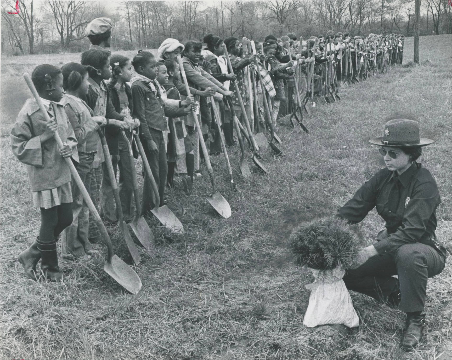 Black and white photo of a line of thirty mostly Black girls, each holding a shovel, with a uniformed woman to the right holding a white bag of pine tree saplings.