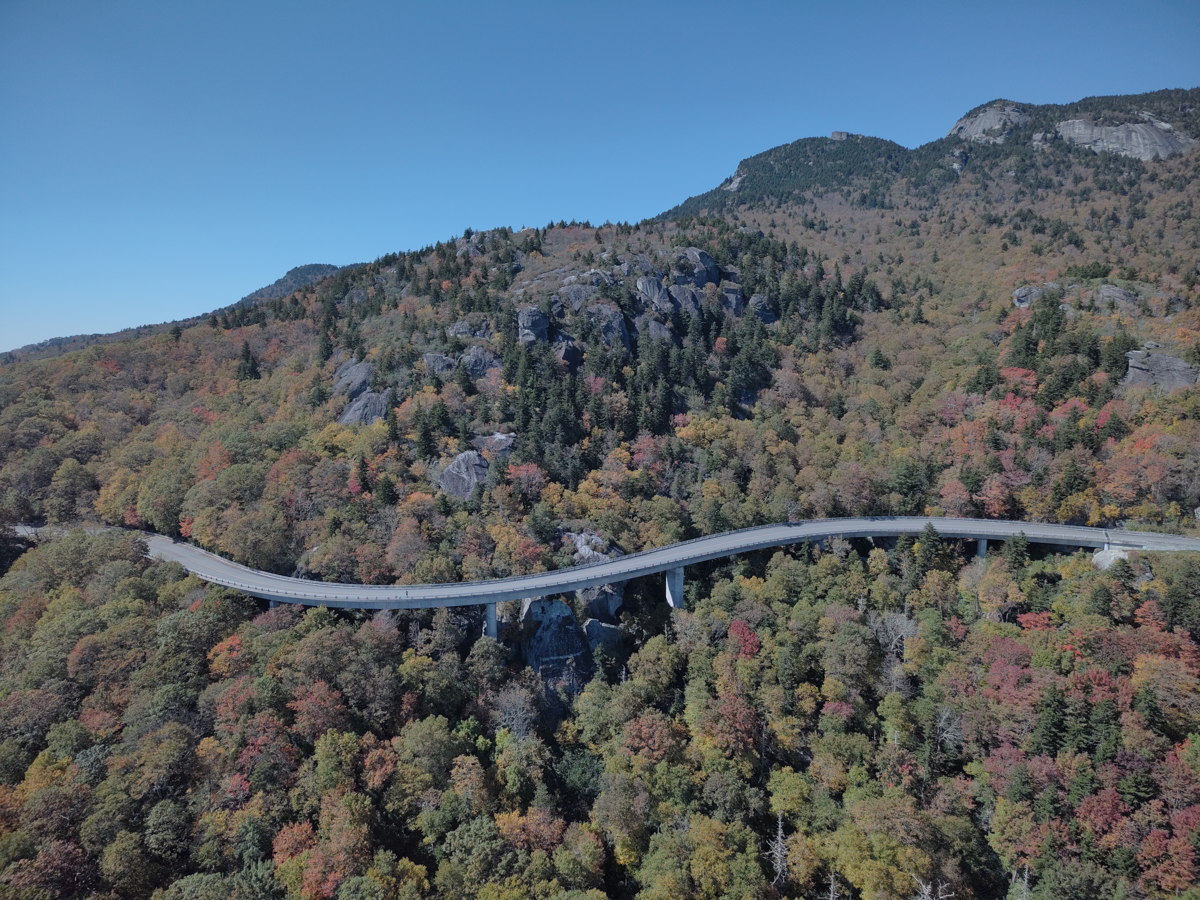 A long, S-curved bridge hugs the side of a mountain. The bridge is surrounded by trees beginning to turn orange and red.