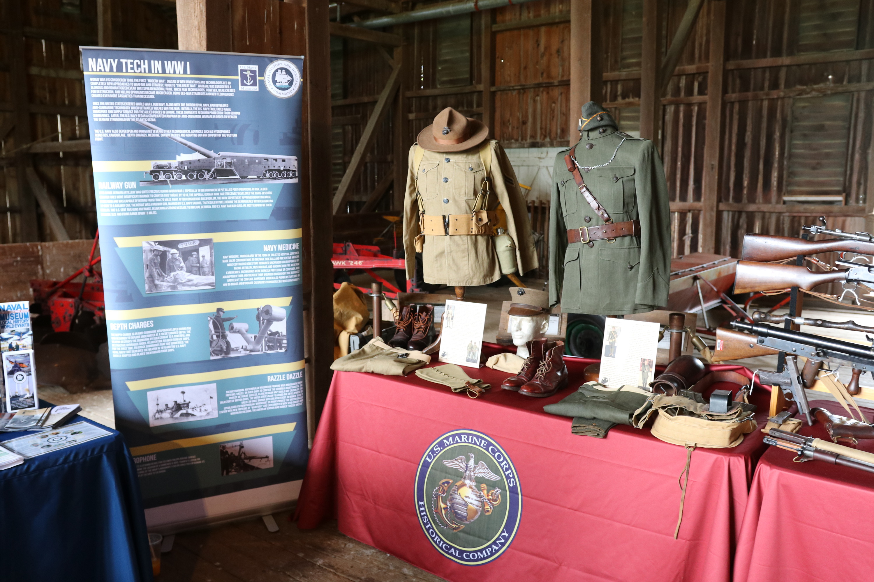A marine corps table display is set up. A large poster of technology utilized in the war stands next to a display table with historical weapons and soldier uniforms.