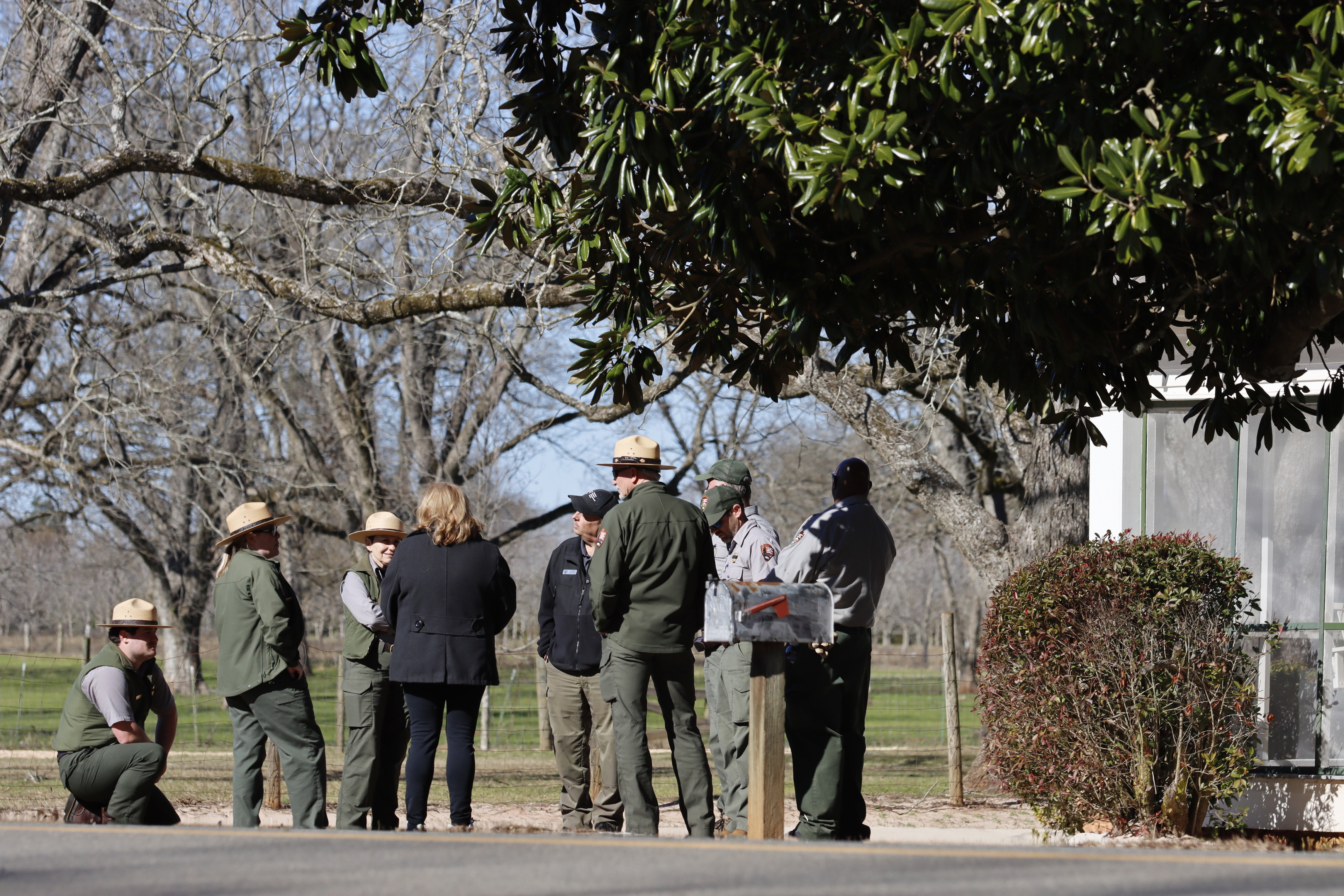 A group of National Park Service employees gathers outside the Boyhood Home for a meeting. Dressed in NPS uniforms, the team stands in a circle, engaged in discussion. The historic Boyhood Home is in the background and the branches of a large tree are above them. 