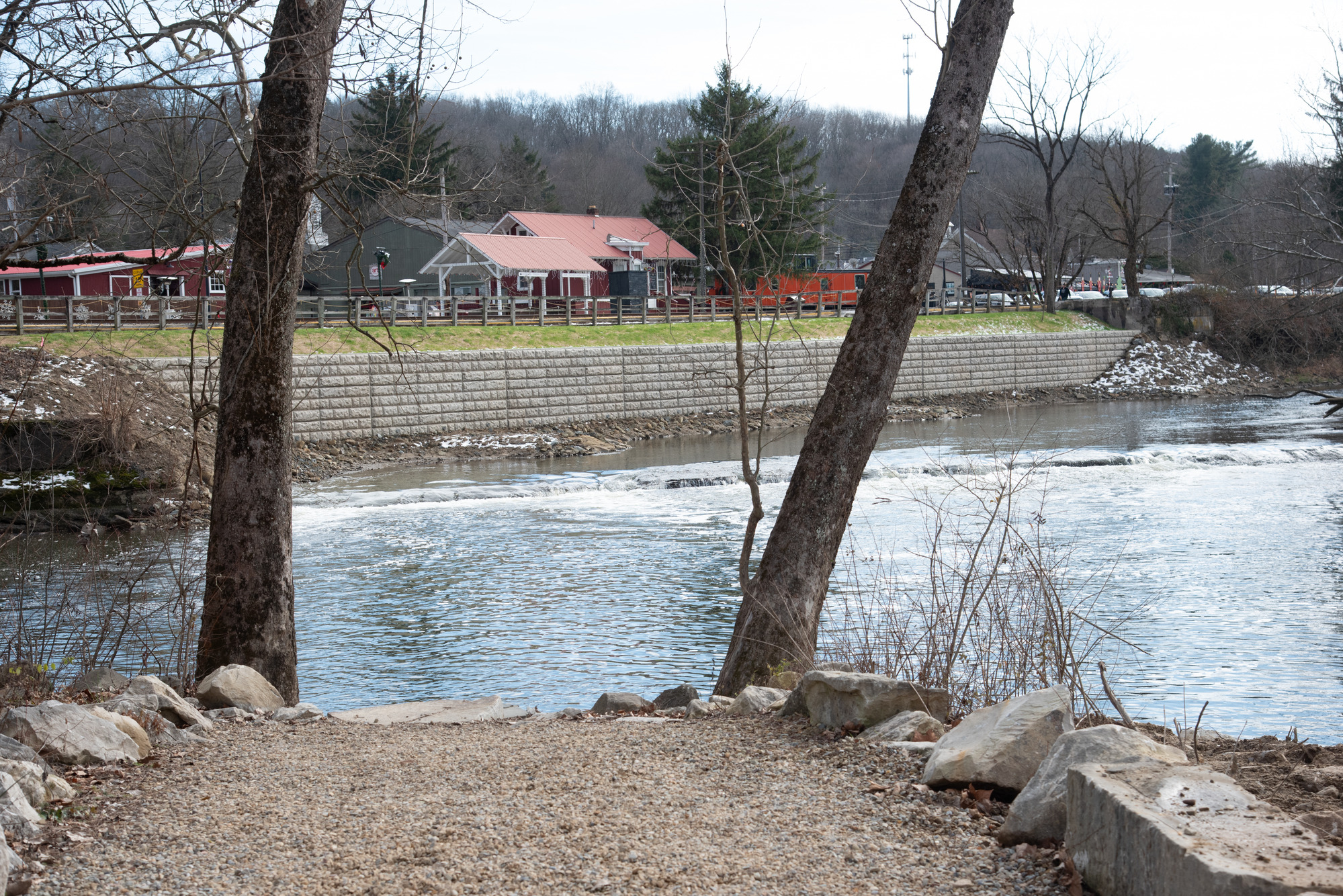A gravel path approaches a body of water; on the far side, a concrete wall lines the riverbank below several buildings.