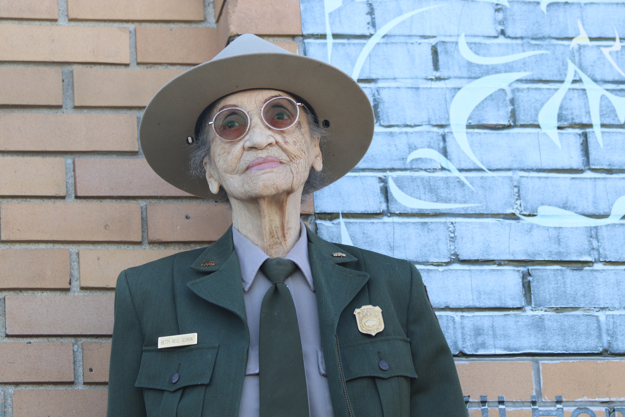 A senior African American woman in a ranger uniform leans against a brick wall and looks up at sky. 