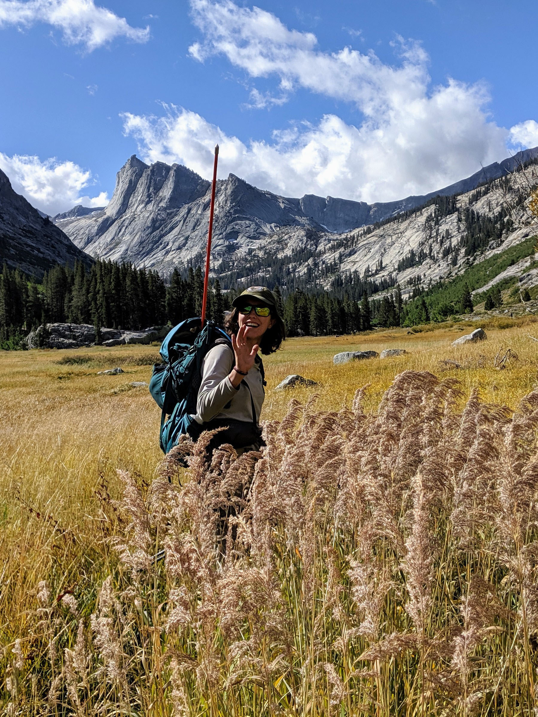 Woman stands in a scenic meadow and carries a backpack and large sampling tool to monitor wetlands.