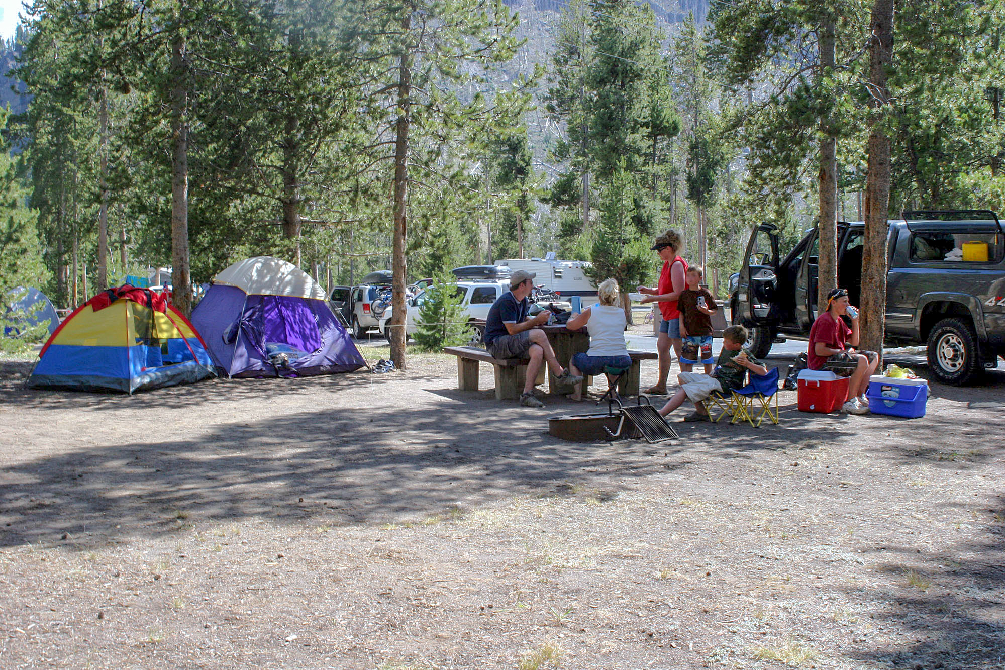 Campers are sitting at a picnic table with two tents nearby.