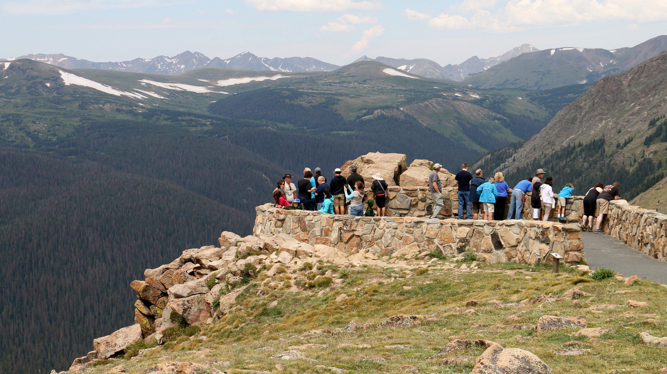 visitors stand in a stone circular lookout point surrounded by snowy mountains 