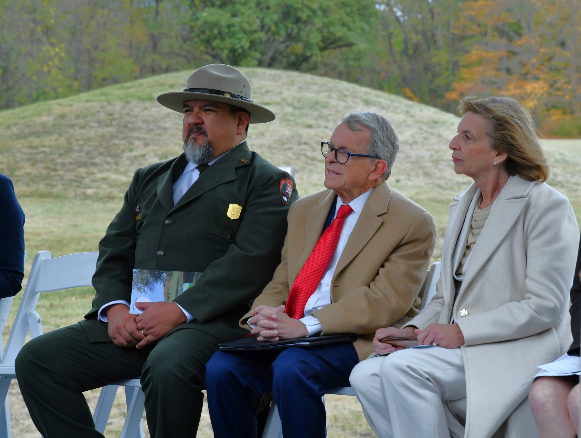 A man wearing a green suit with green tie and tan, flat-brimmed ranger hat sits to the left, a man in a beige coat with red tie and white shirt sits in the middle and a lady wearing a white coat with light-colored blouse sitting on the right.