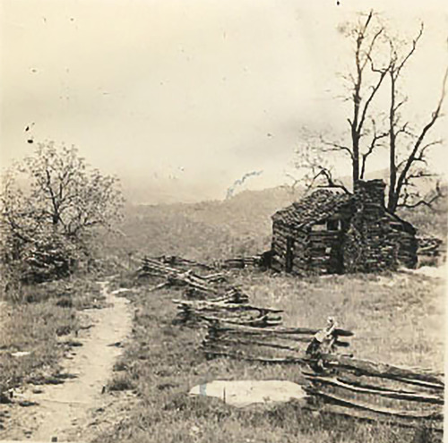 A black and white photo of a small log cabin behind a split rail fence. Two leafless trees are behind the cabin and a rough dirt path runs in front. 