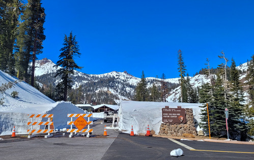 A photo of a mountain road cleared of snow. A white truck is parked next to a cut snowbank behind a road closed sign.