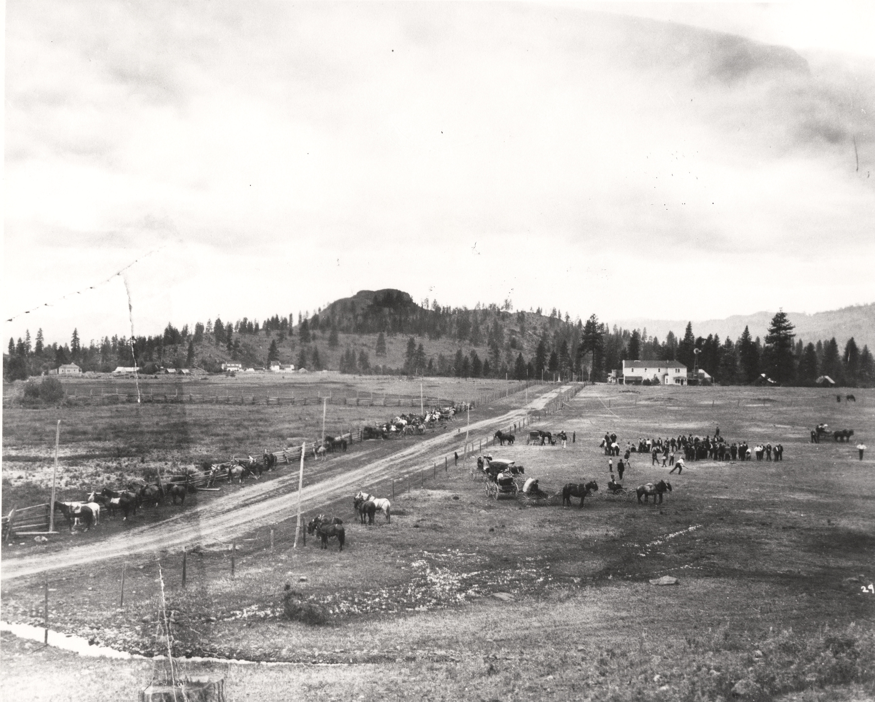 Black and white photograph of a road through an open field with people, horses, and buggies gathered next to the road. 