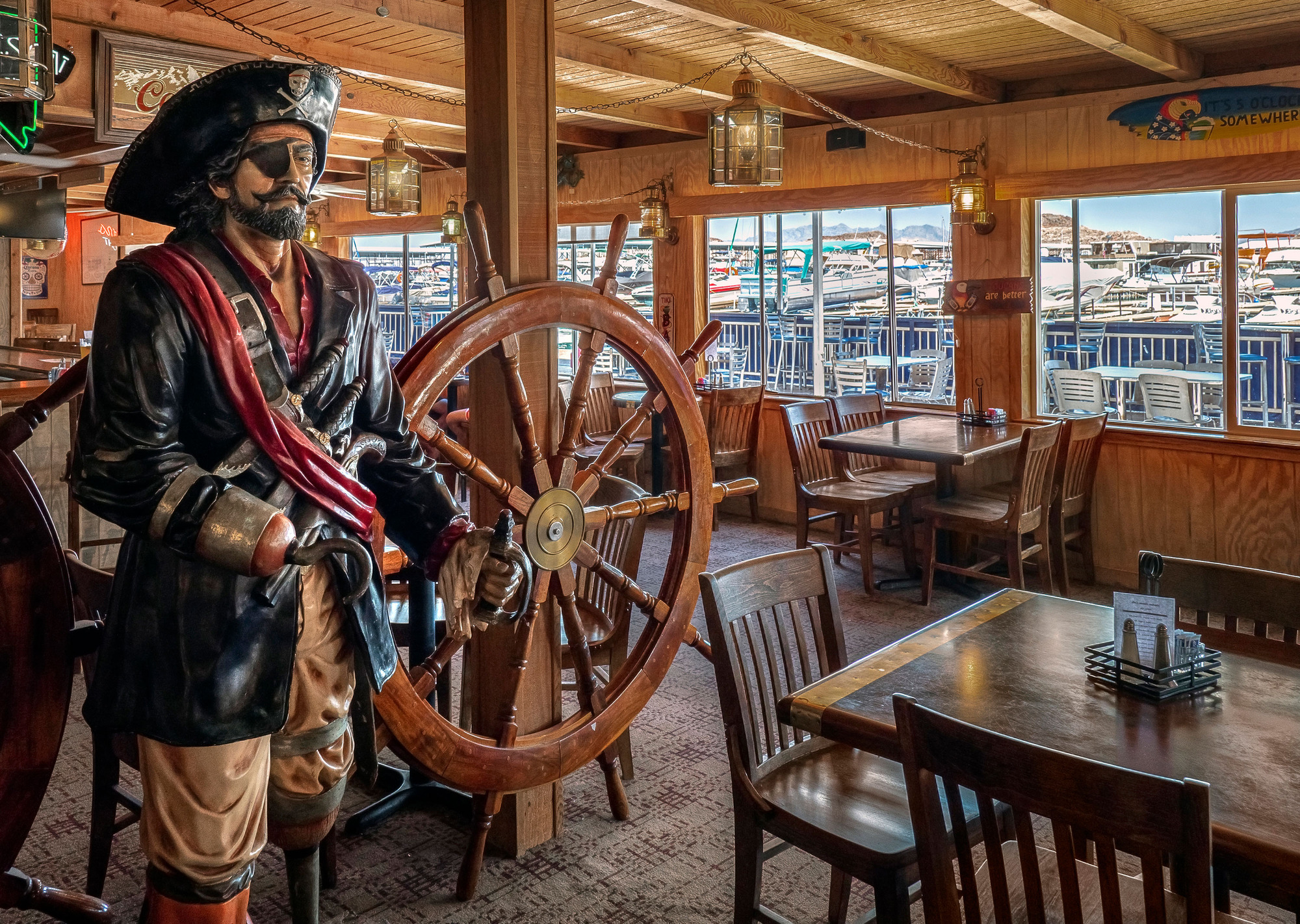 Stature of pirate and ship's wheel on left, empty tables in restaurant, marina seen outside 
