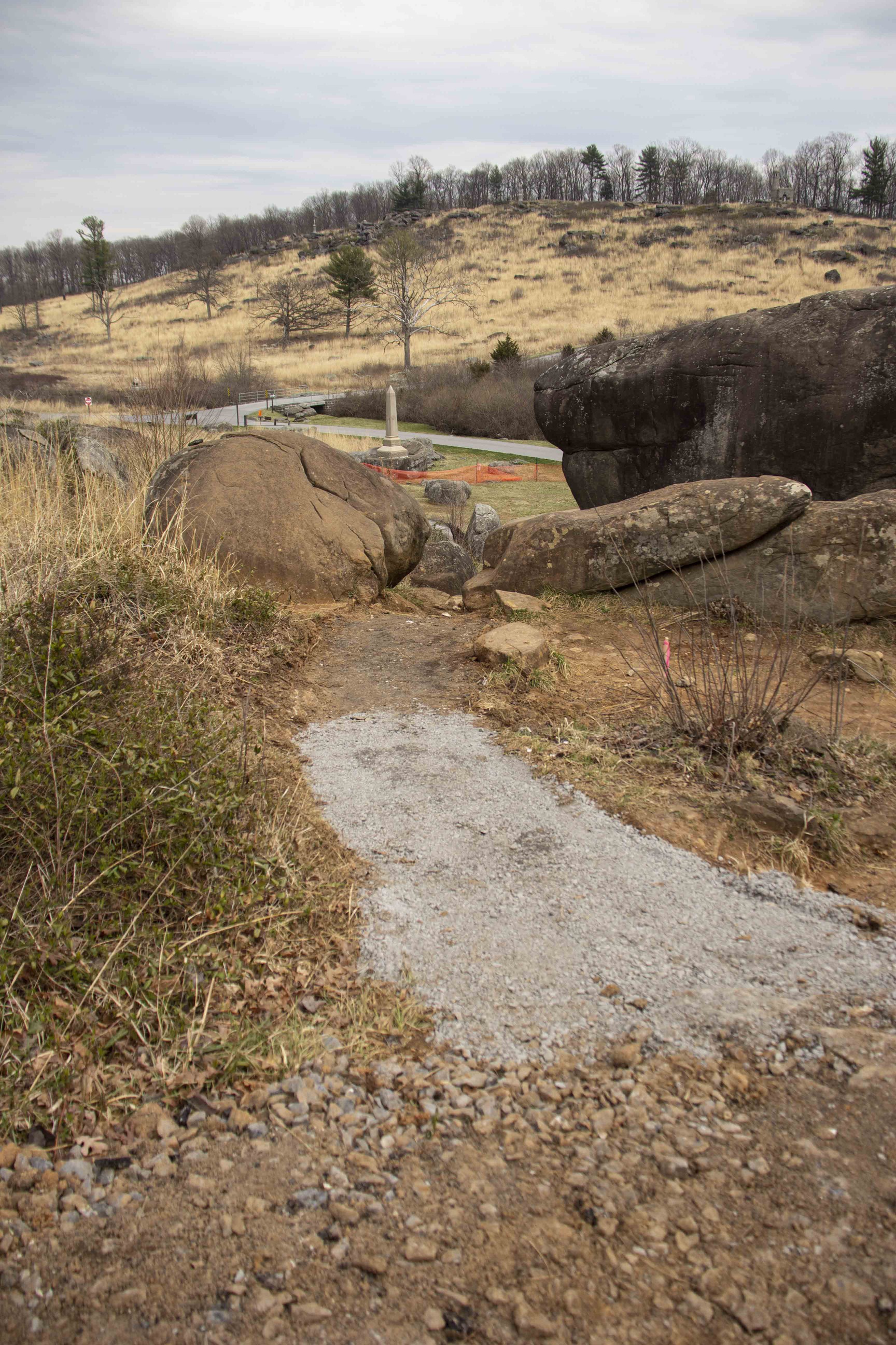 The focus of the frame is a dirt path with its pavement recently removed leading vertically up the frame and down a slope. There is gravel visible on the path near the camera. There are large boulders on either shoulder of the path up ahead. In the far distance down the slope is a stone obelisk surrounded by orange construction fencing.