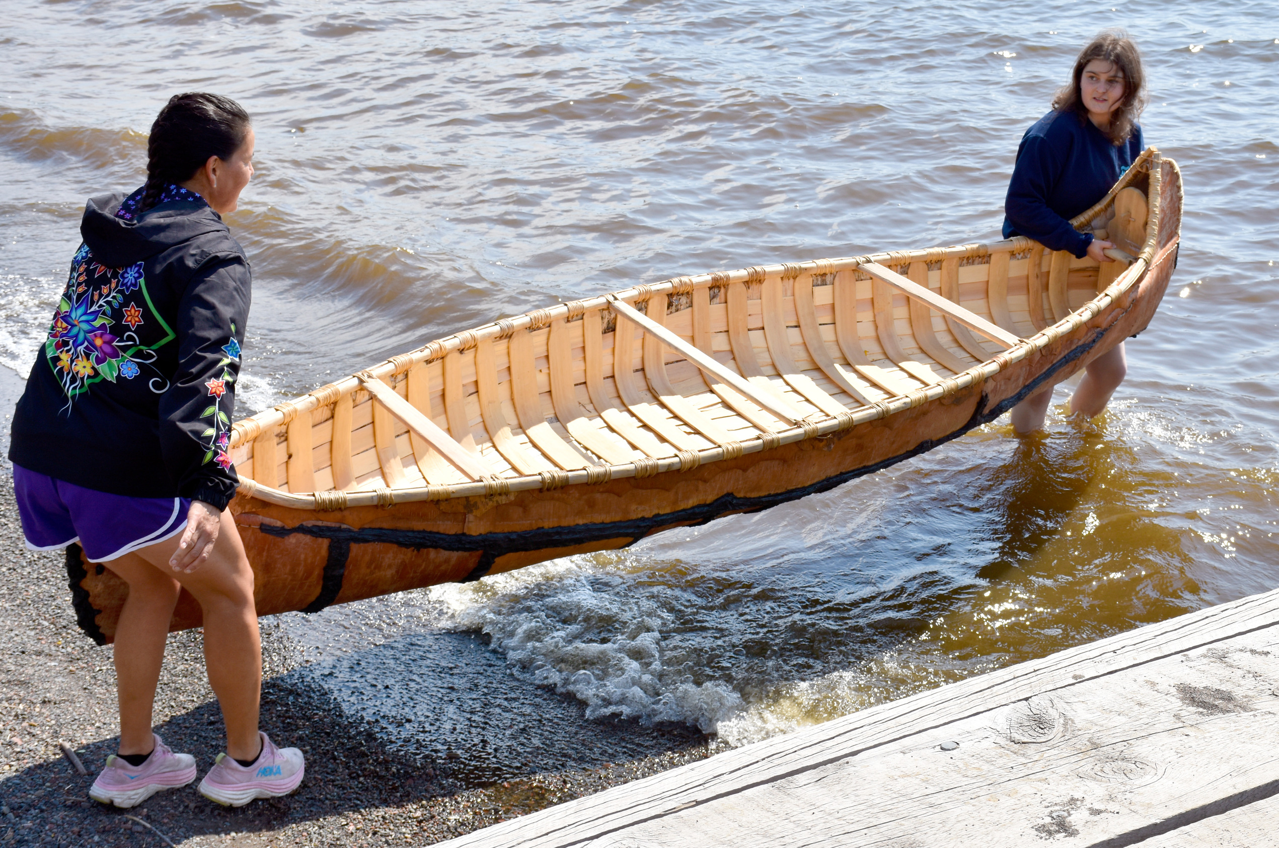 Two people hold a wooden canoe over water at the shoreline.