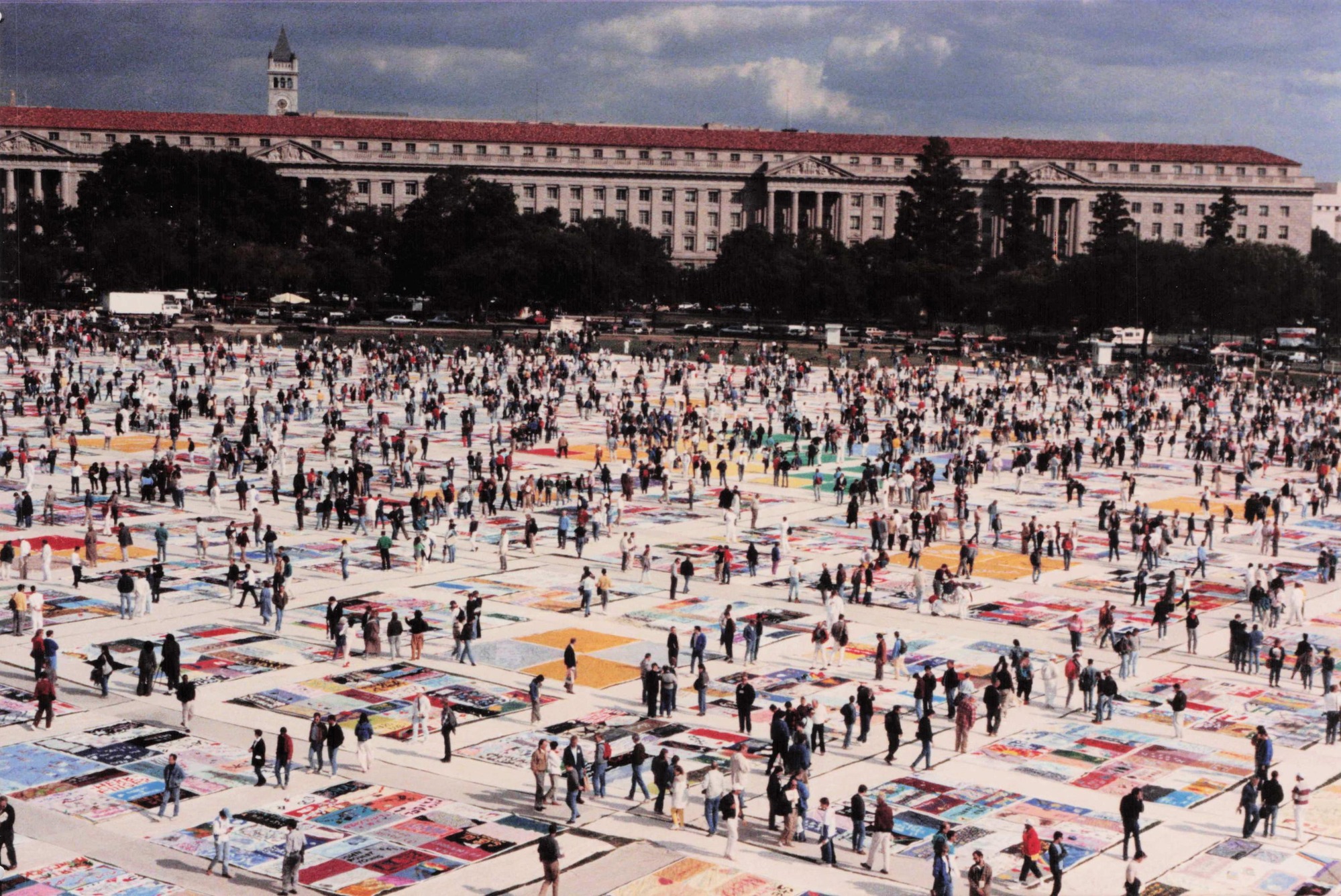 Multi-colored panels are laid on the grass. Dozens of people walk on top of the panels and stand on the side. A building and tower are visible in the distance