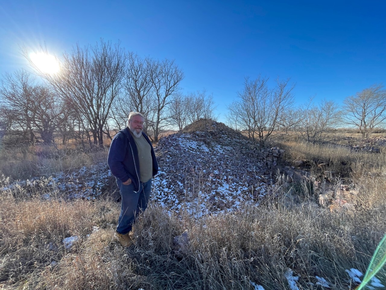 A man stands next to a pile of rocks in front of trees on a sunny day.