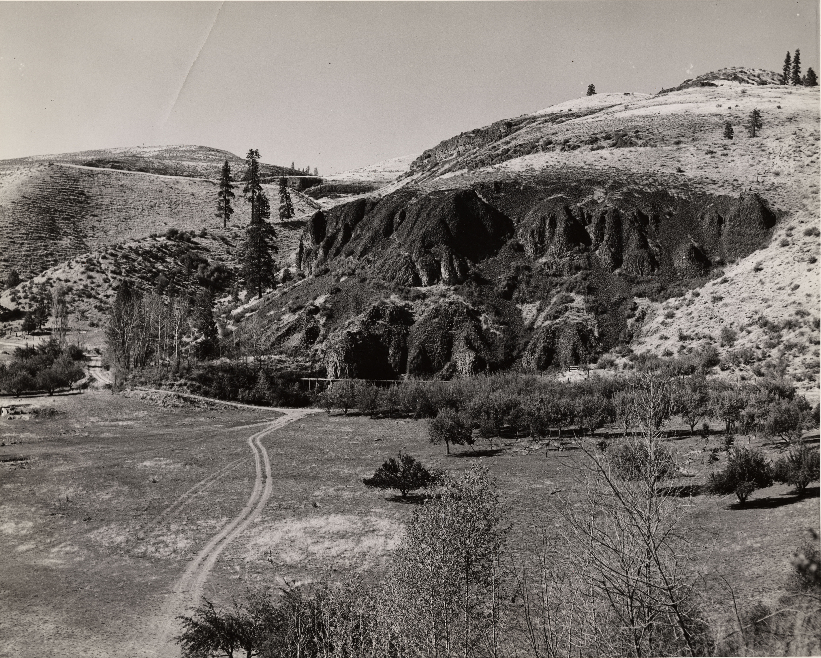 Black and white photograph of a large clearing with a dirt road across it with steep, dark cliffs in the background