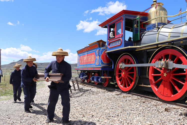 Four men dressed as Chinese immigrant railroad workers from the 1860s carry and iron rail alongside the red and blue steam locomotive Jupiter.
