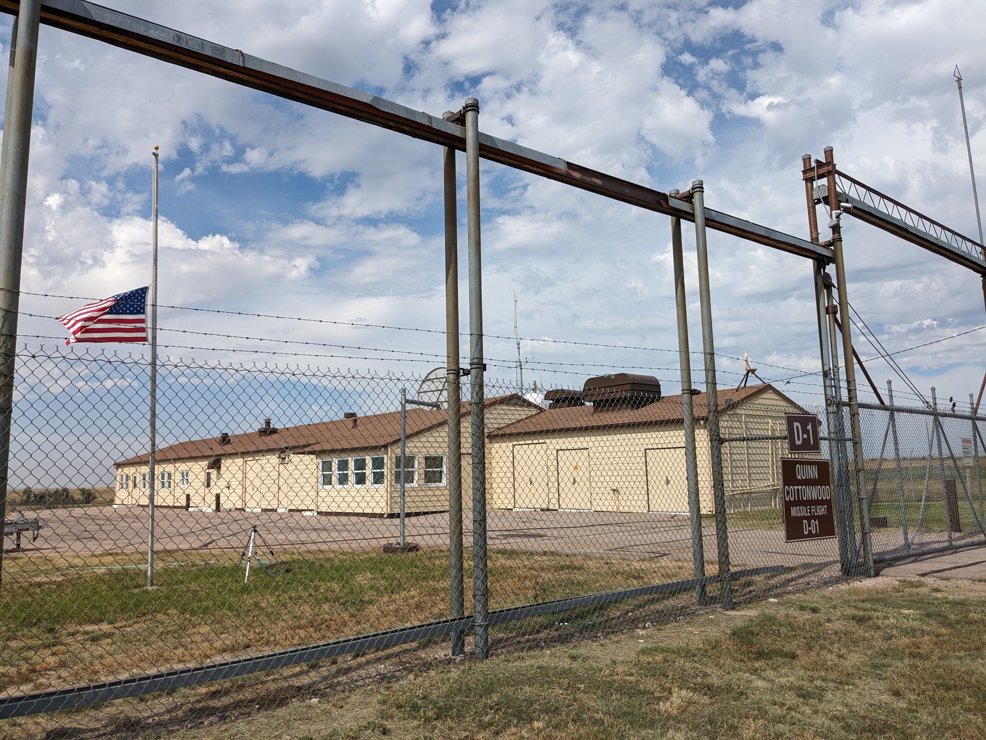 Long beige building behind a chain link fence. The flag on the pole in front of it is flying at half mast. 