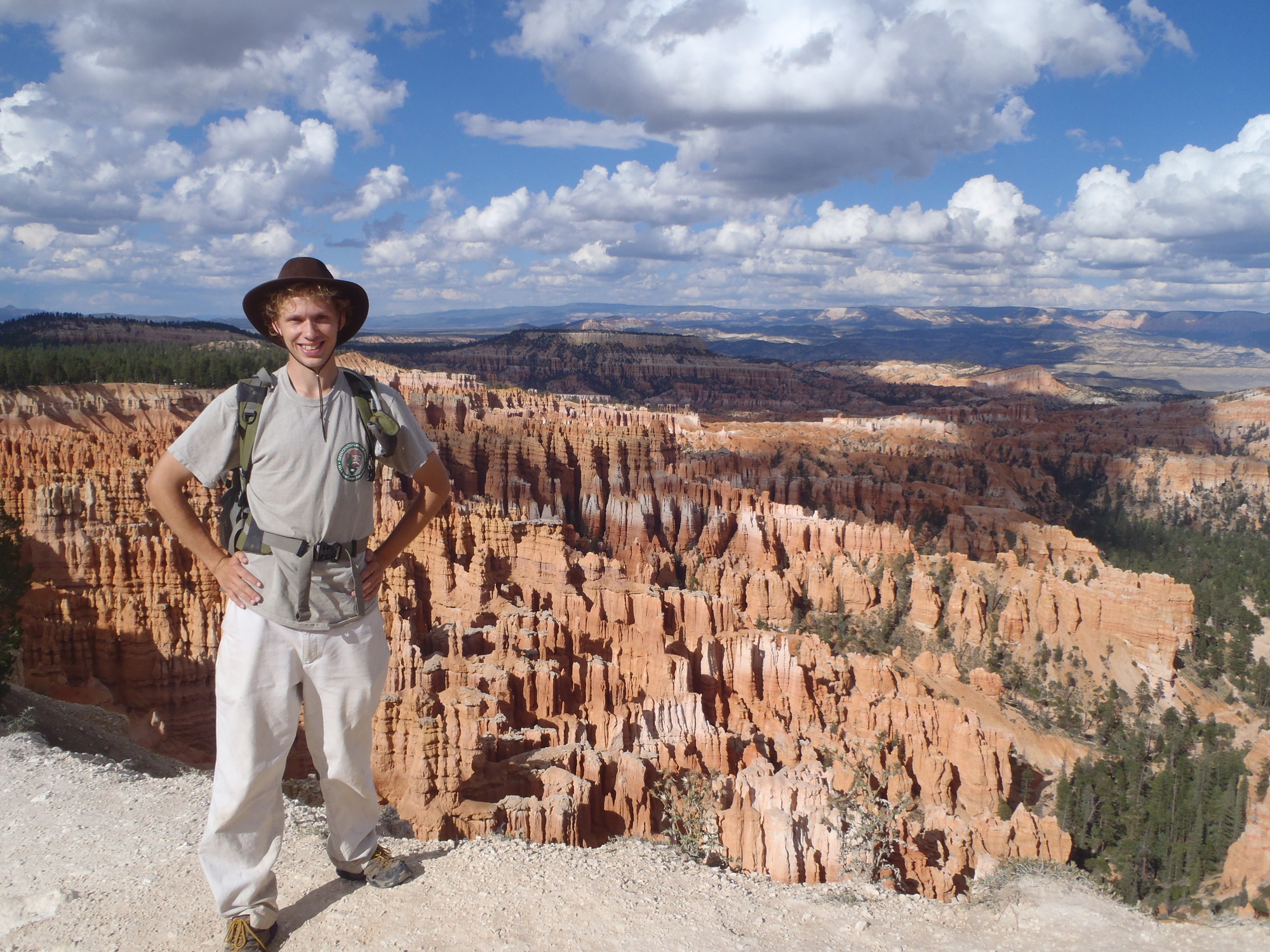 a young man in a wide brimmed hat stands on the edge with tall spires of rock and blue sky in the background.