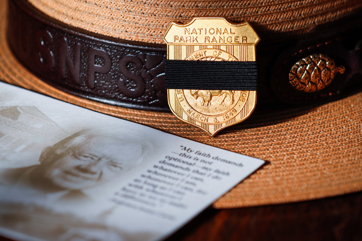 A Park Ranger's flat hat rests on a surface, prominently displaying the National Park Service badge on the front. A black mourning band wraps around the badge. The memorial card for Jimmy Carter sits overlapping the hat reads, "My faith demands—this is not optional—my faith demands that I do whatever I can, wherever I am, whenever I can, for as long as I can with whatever I have to try to make a difference.” -President Jimmy Carter