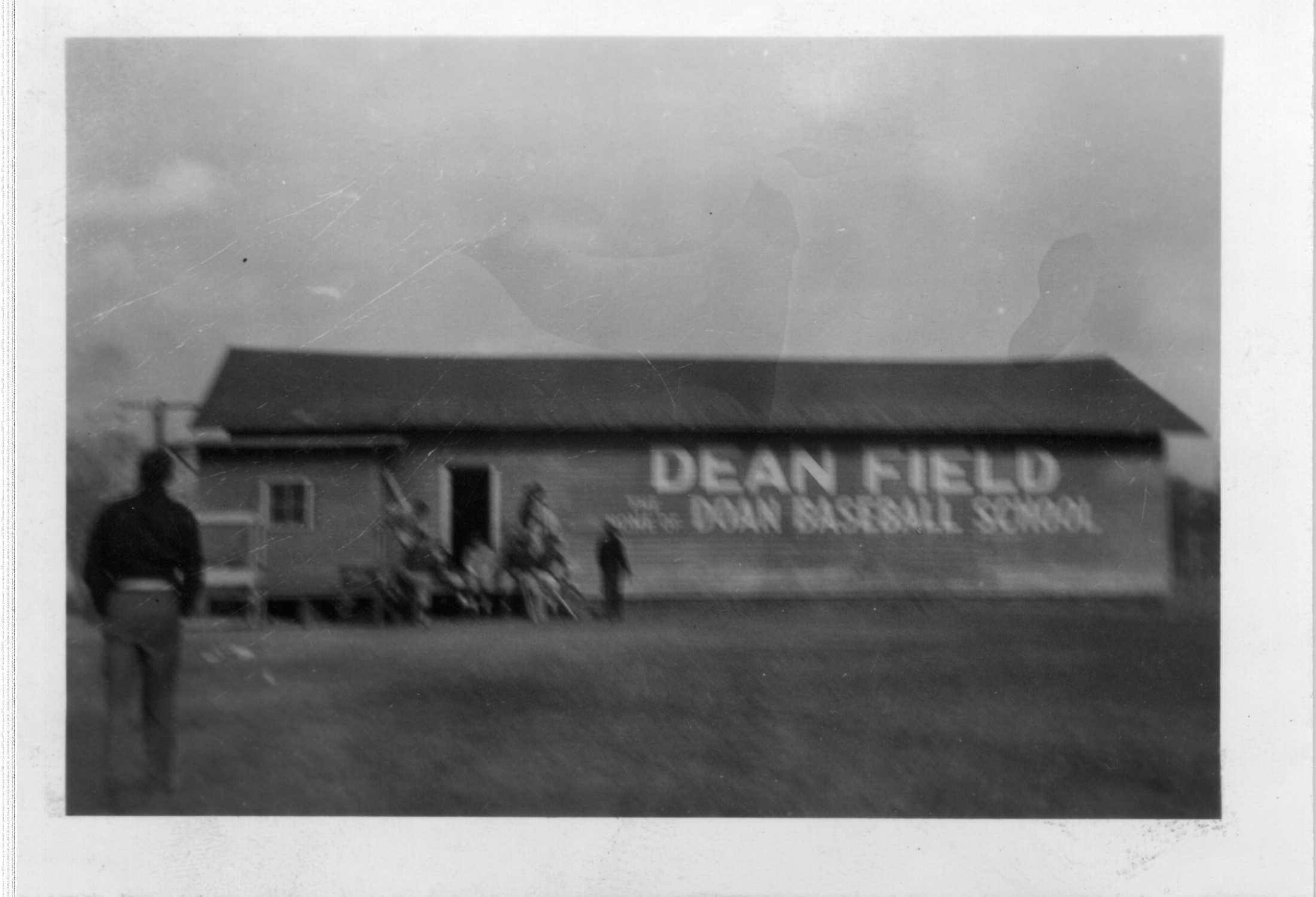 Small black and white photographic print of Dizzy Dean's baseball school in Hot Springs, Arkansas.
"DEAN FIELD" "DEAN BASEBALL SCHOOL," painted in white paint on the wall of a long, possibly unpainted wood frame structure with 1 door and 1 window. Small group of players gathered around door; in left corner of foreground, man with his back to camera is walking toward building.