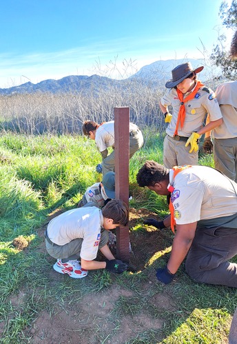 6 people working together to install a post amidst grass. Mountains soar in the background.