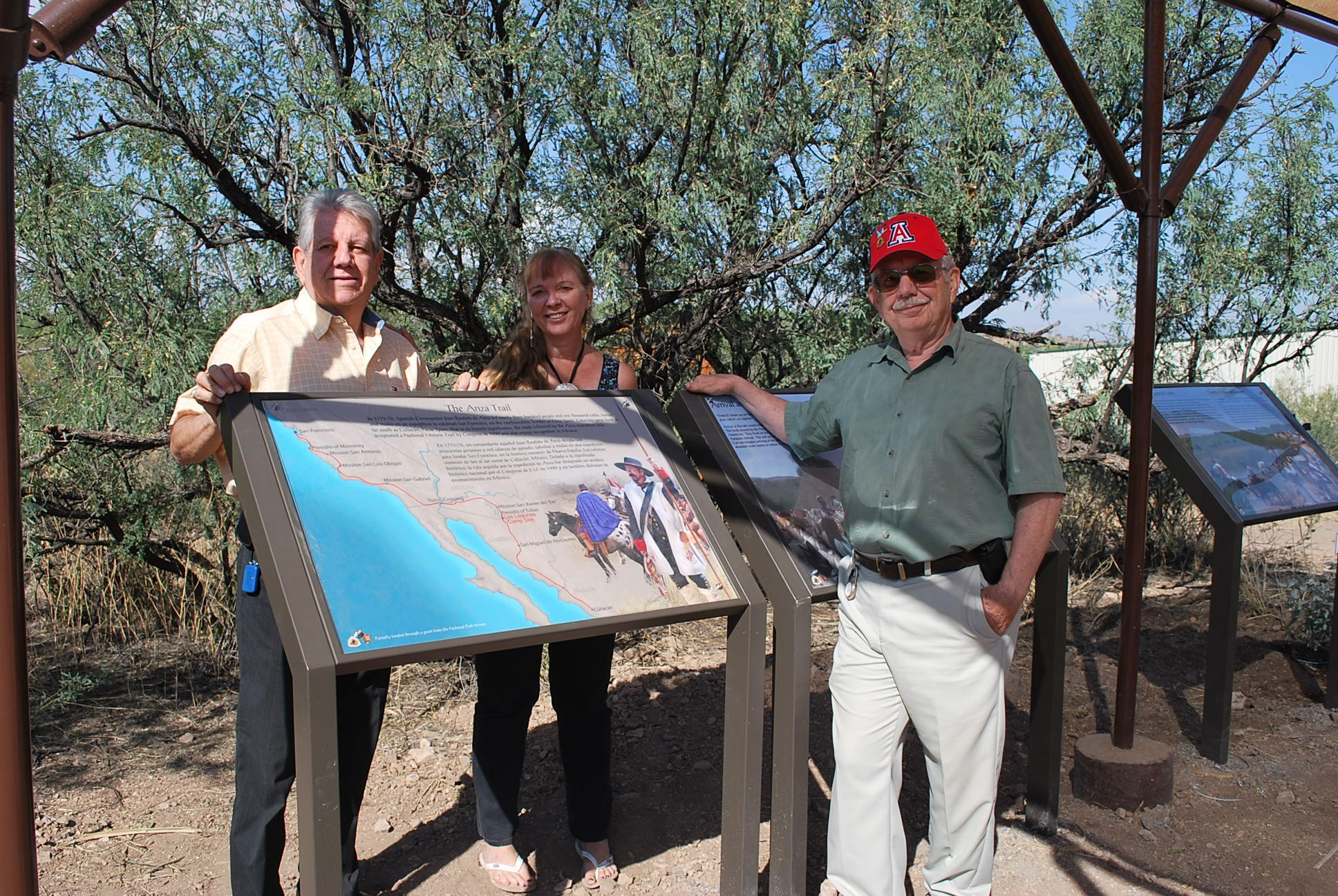 Three people gather around one of three interpretive panels outside under a shaded structure