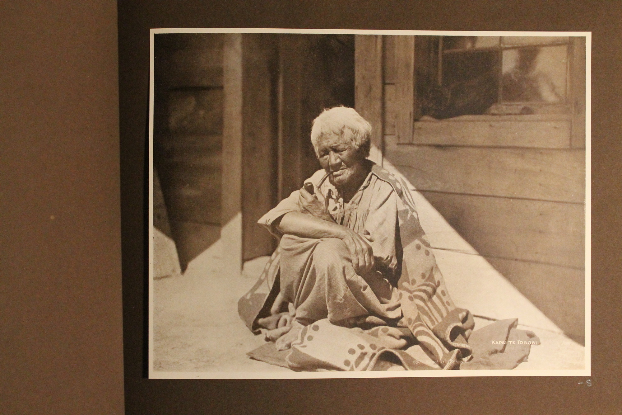 An elderly Maori sits on her heels smoking a pipe in front of the doorway to a wooden house.