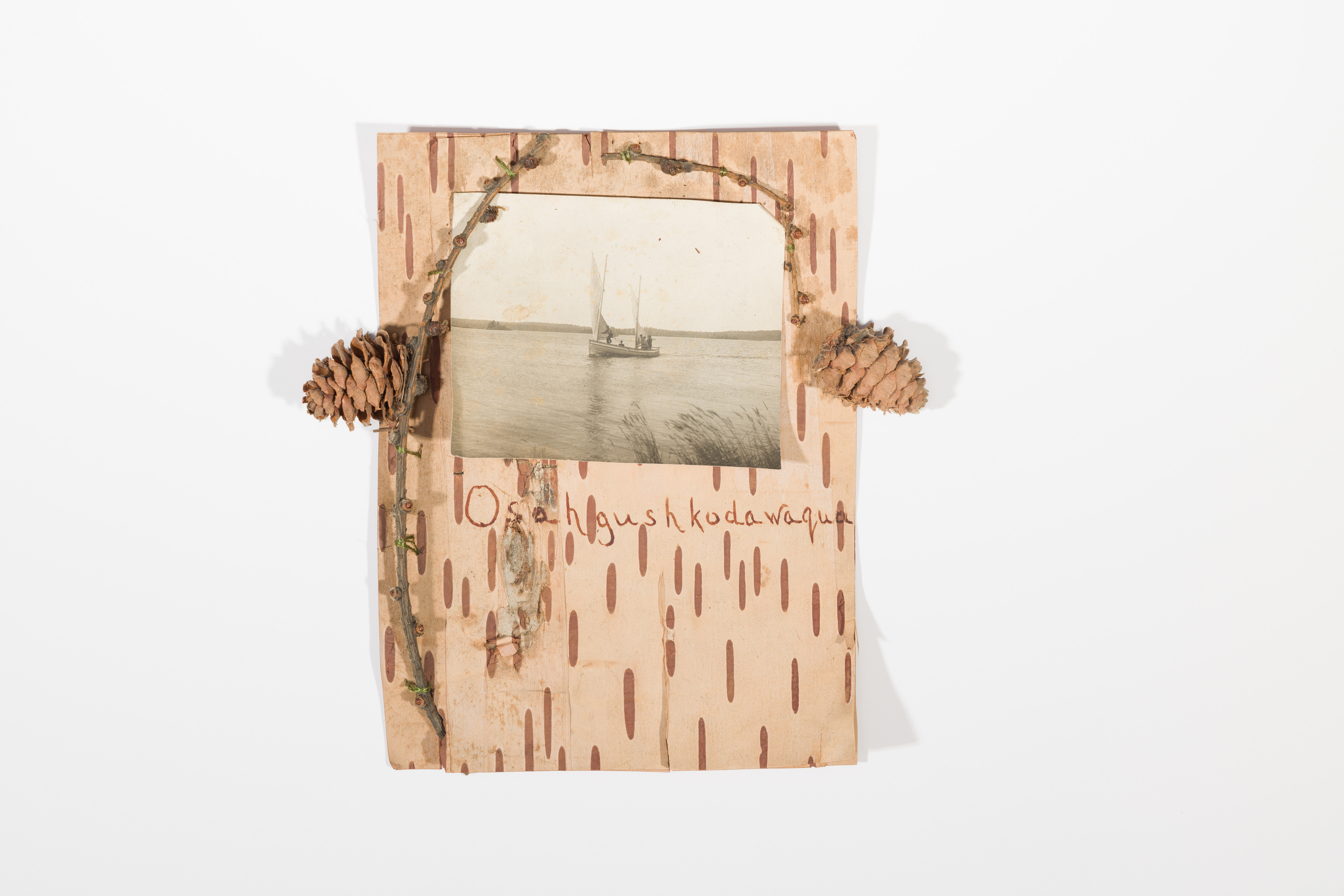 A piece of birchbark with three twigs attached by green thread framing a photo of several men on a boat.