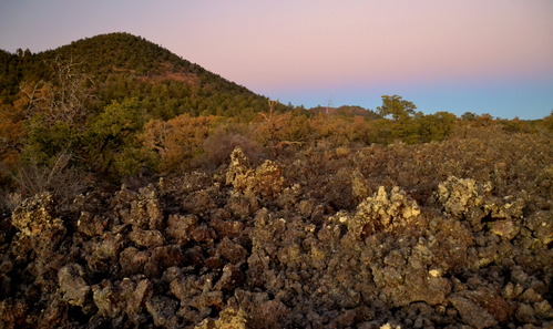 A rounded, conical peak stands behind a jagged line of black rock and shrubs.