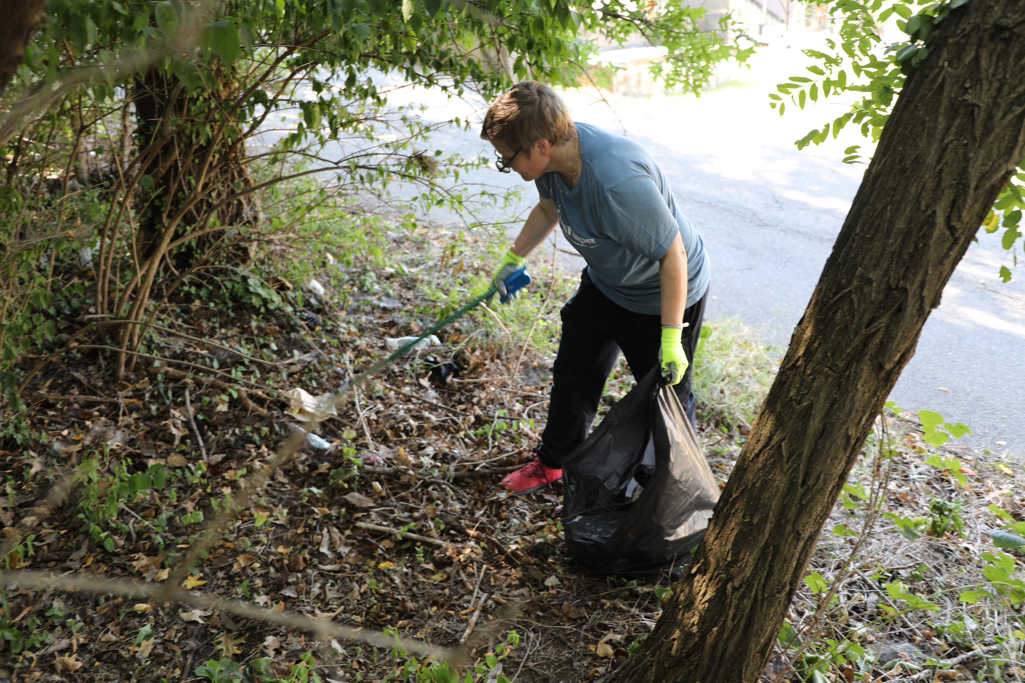 A person wearing a blue t-shirt, dark pants, and bright yellow-green gloves is picking up trash from a brushy, overgrown area. They are holding a black trash bag open with one hand and are using a trash picker tool with the other to grab a piece of litter. In the background, a paved road is partially visible.