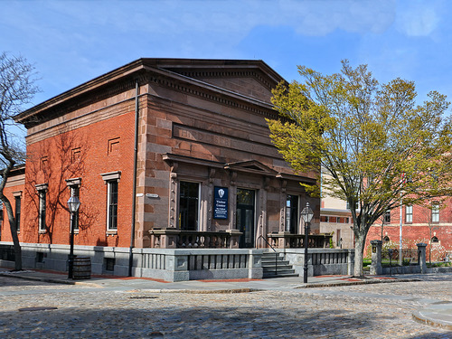 New Bedford Whaling National Historical Park Visitor Center on a bright fall day. Looking diagonally across the cobblestone streets you will see the left side of the building with three windows and red brick. To the right is the varied pink granite along the front side of the building with two windows and a double door down the center, with grey stone steps leading to it.  