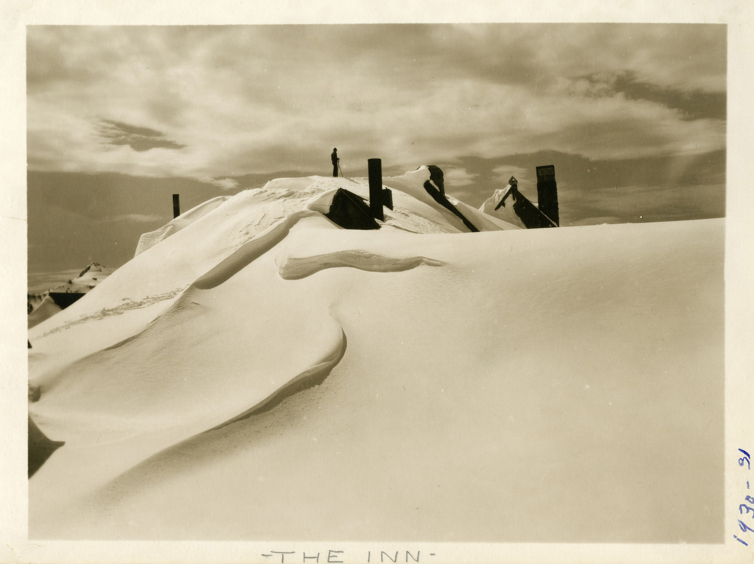 Historic photo of a building almost completely buried under snow with just the fireplaces sticking out and the shape of the snow revealing an angled roof. A person stands silhouetted on the snow on top of the building. 